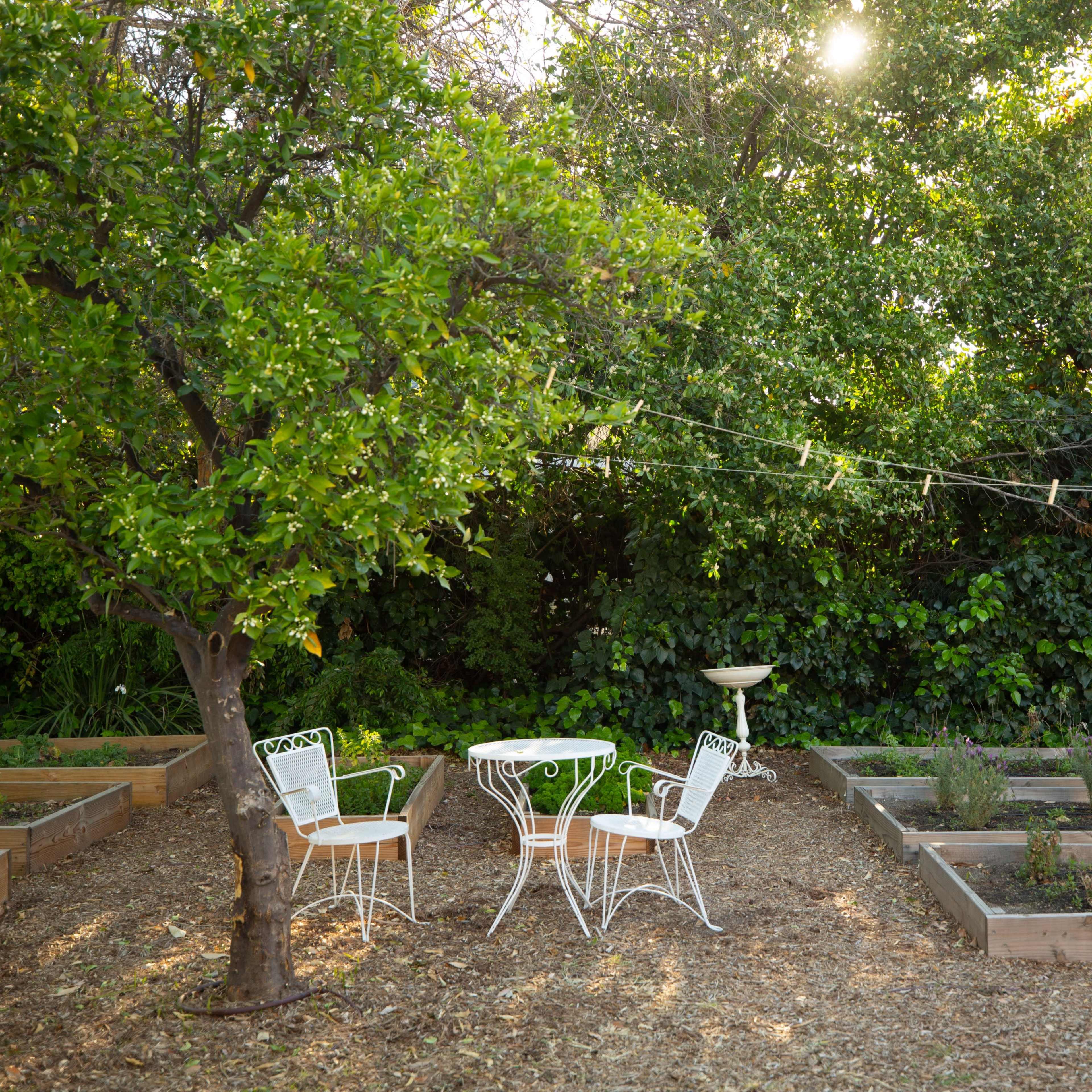 A small outdoor garden features wooden raised beds, a white metal table with two chairs, and a tree providing shade.