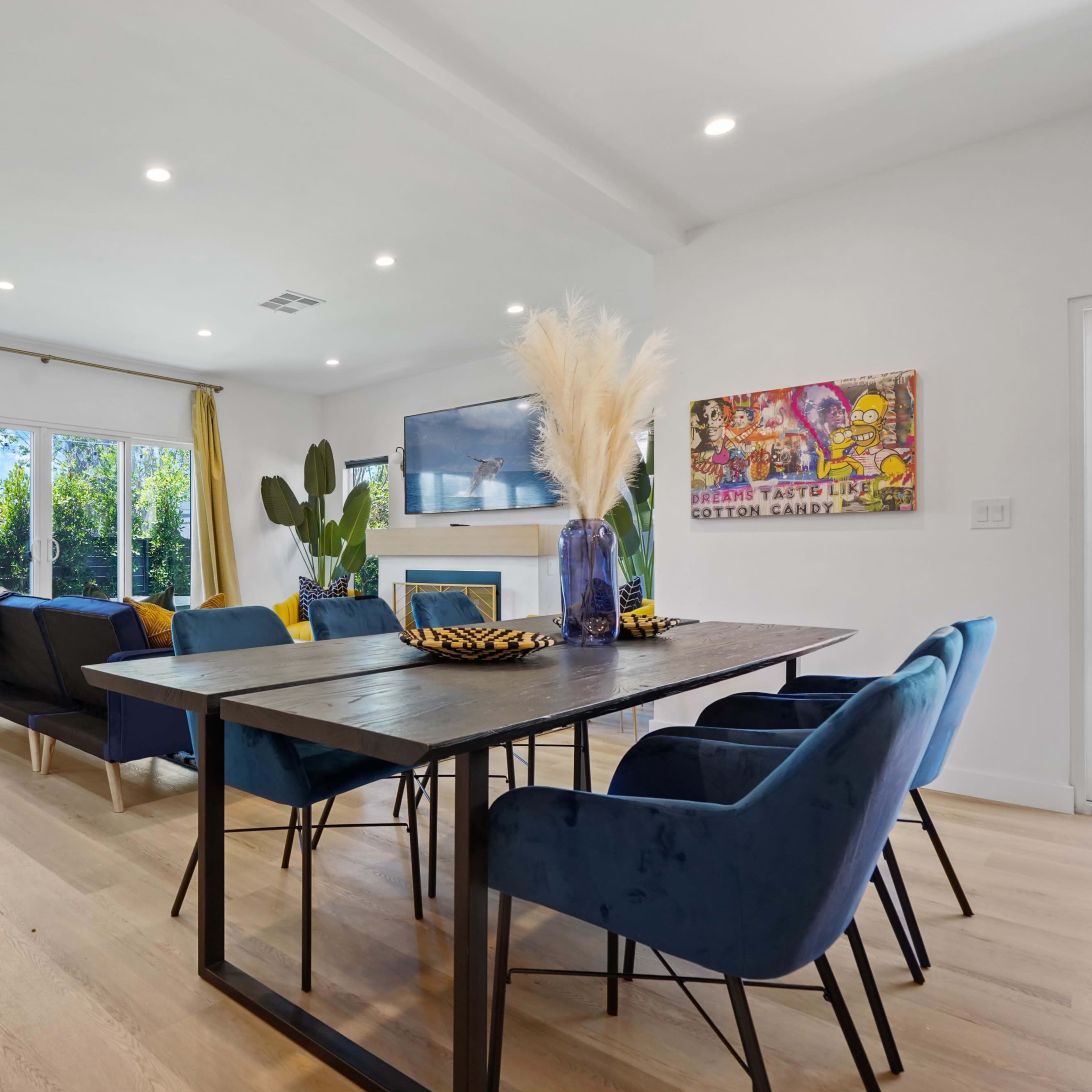A modern living space features a dining table surrounded by blue chairs, a sofa, large windows with yellow curtains, and decorative plants.