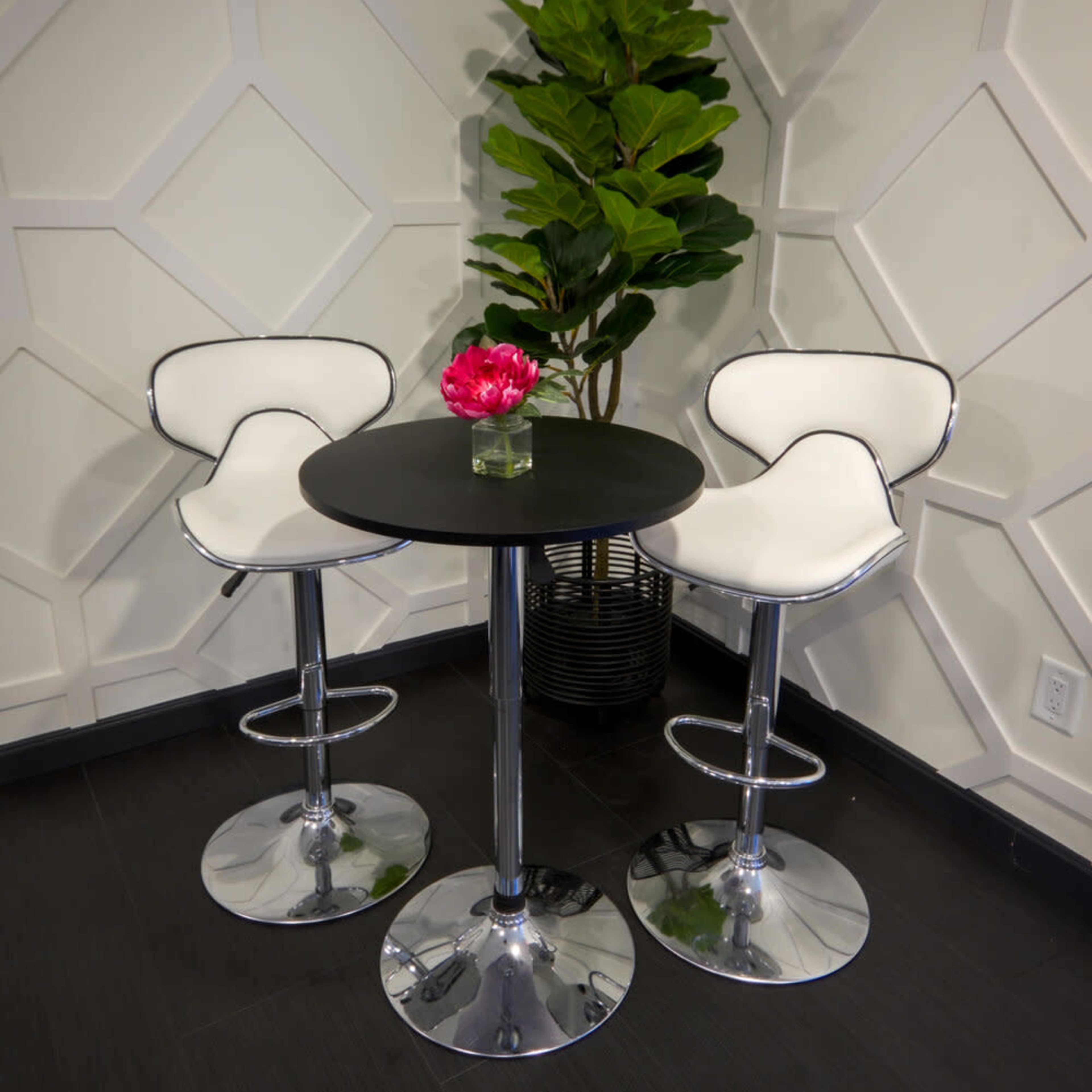A modern black table with two white bar stools and a small pink flower arrangement stands beside a leafy green plant against a textured white wall.