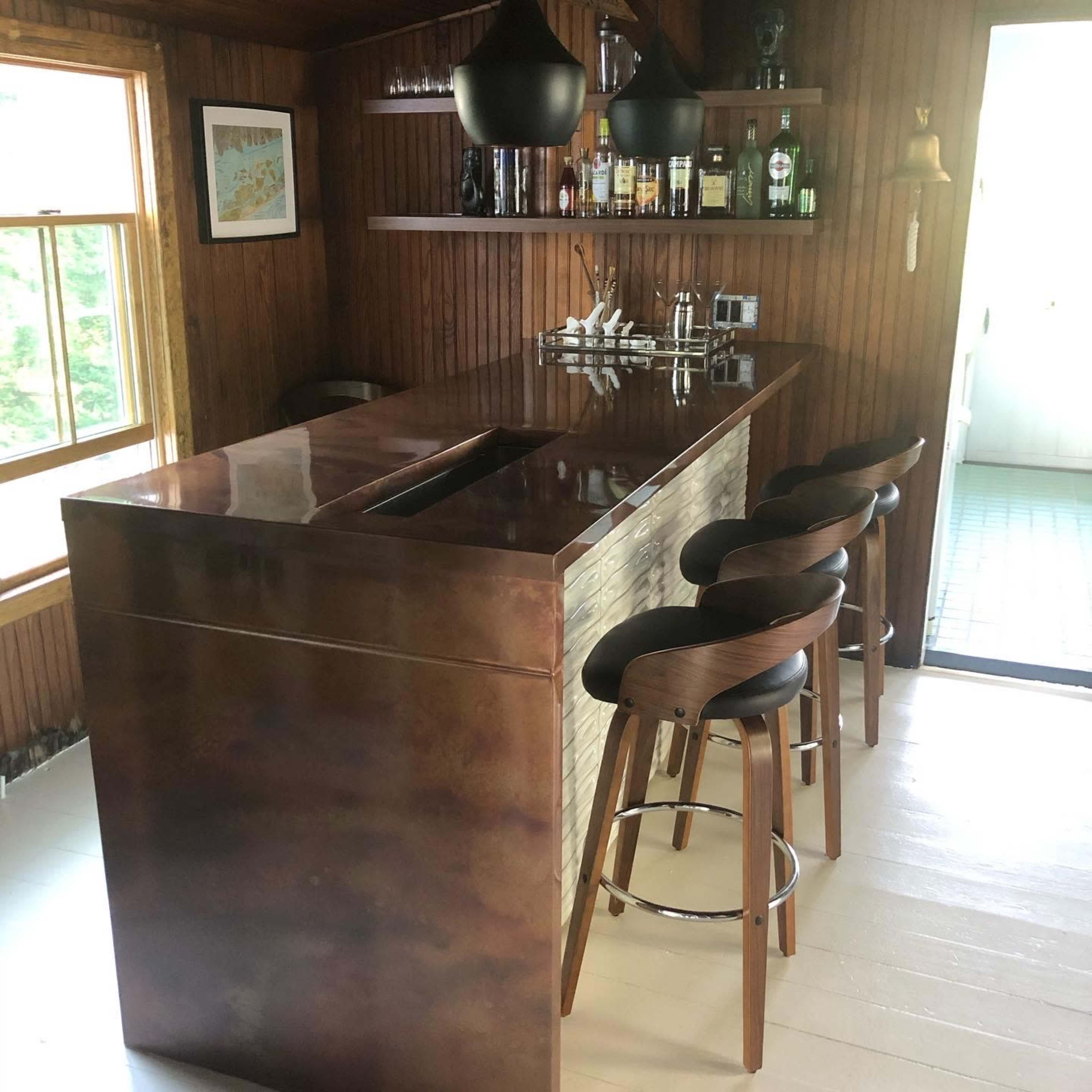 A wooden bar area with a metal countertop, several bottles on shelves, and four high-backed stools.