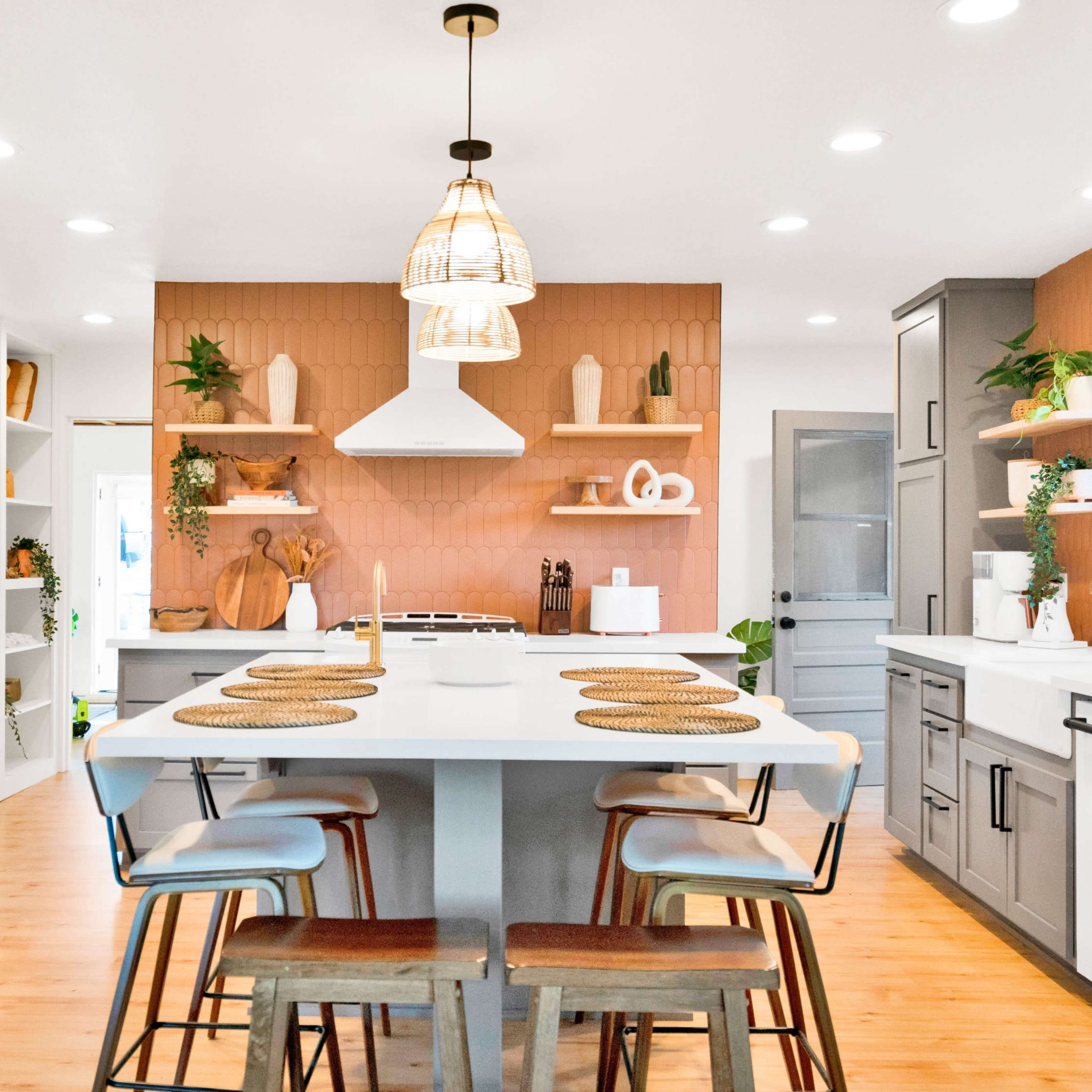 A modern kitchen features gray cabinets, a central island with wooden stools, and warm-toned accent walls adorned with plants and decorative shelves.