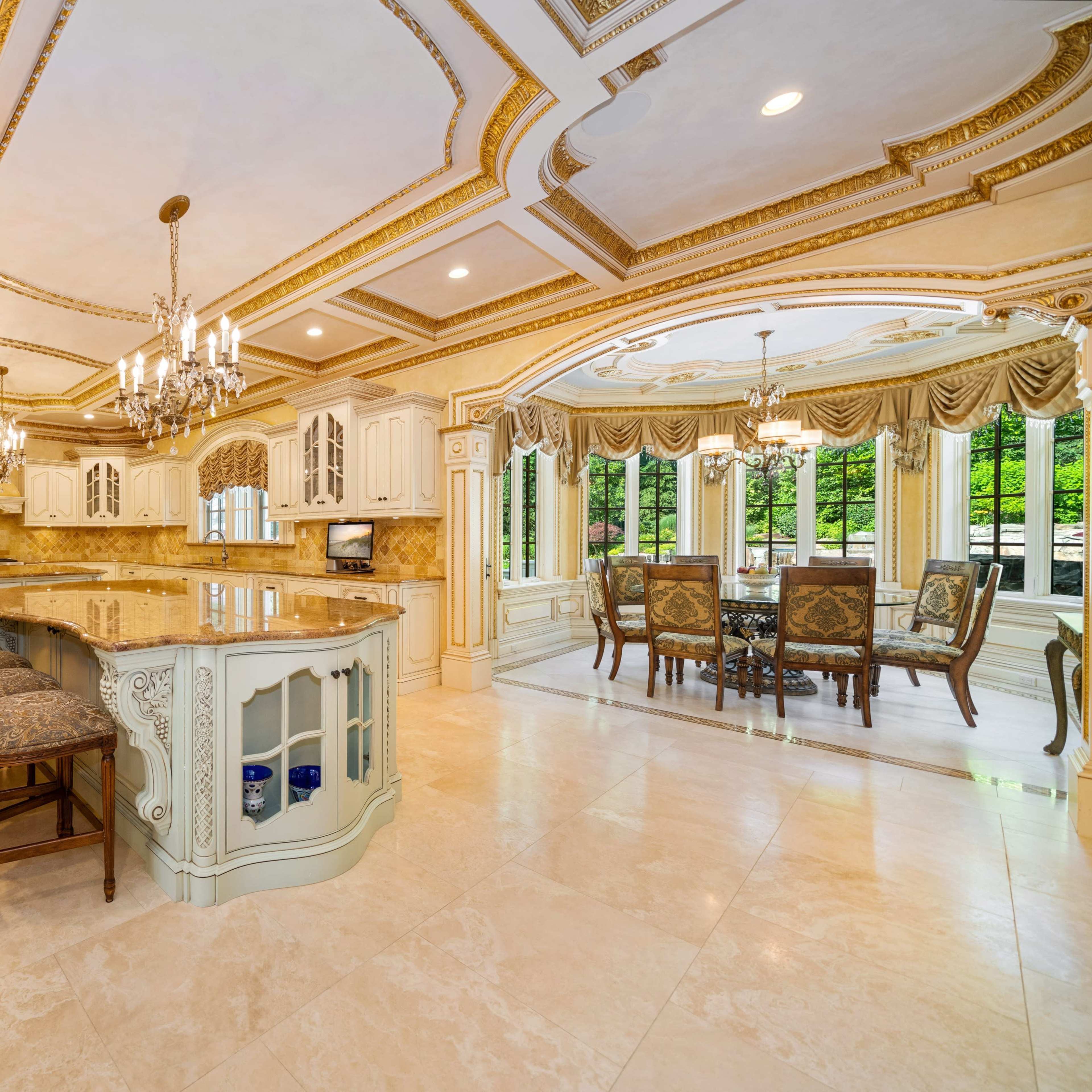 A spacious kitchen with ornate cabinetry, a large island with seating, and an adjoining dining area featuring large windows and elegant furnishings.
