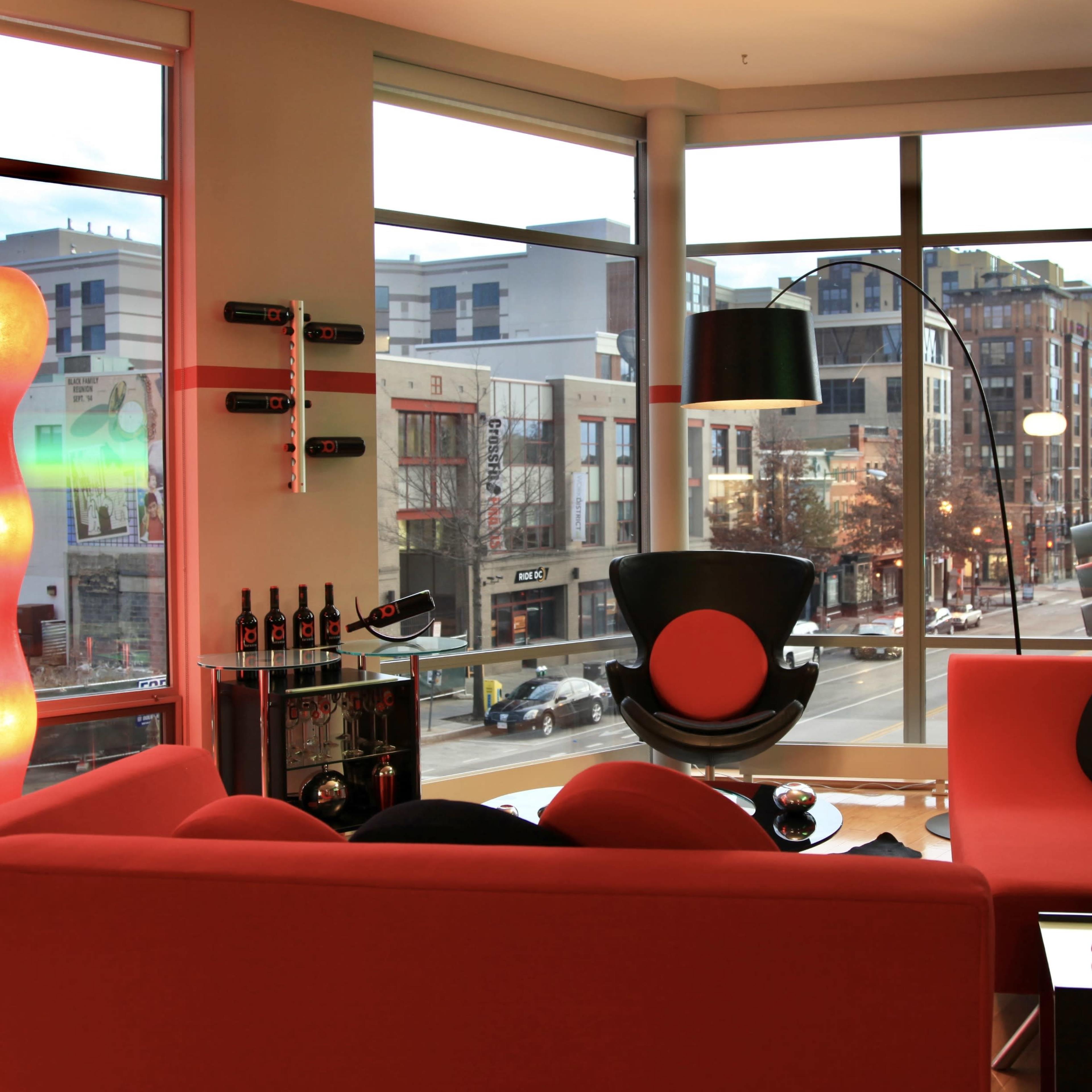 A modern living room with large windows overlooking a street, featuring red furniture and decorative items.