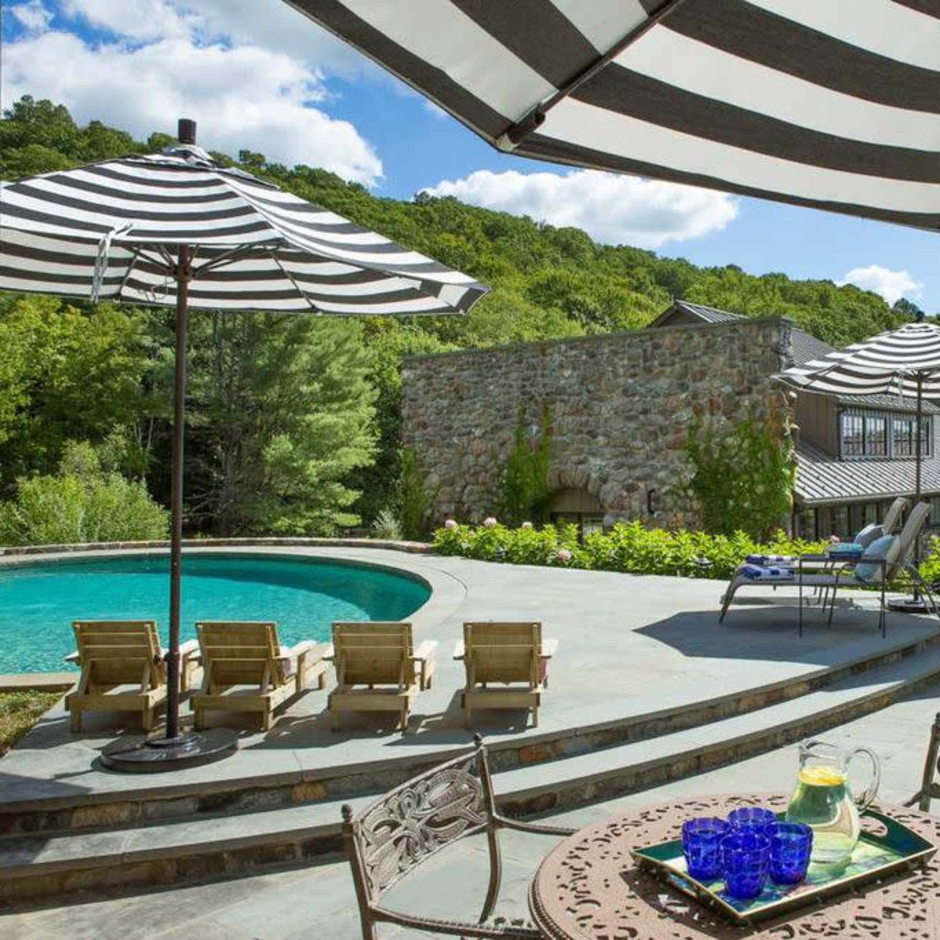 A well-designed outdoor space featuring a pool, lounge chairs, and striped umbrellas, with a stone wall and a house in the background surrounded by greenery.