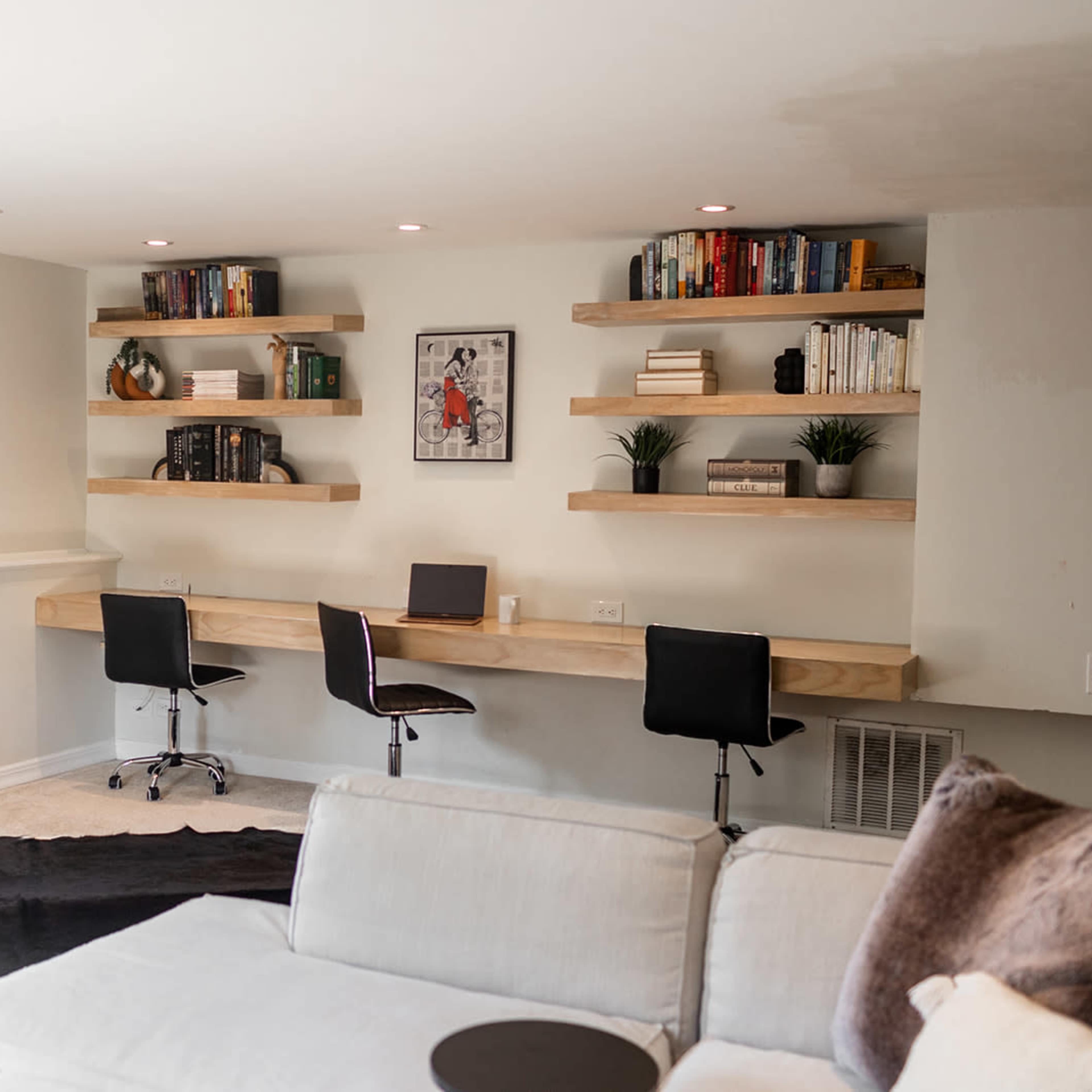 A minimalist home office features a long wooden desk with two chairs, surrounded by wall-mounted shelves filled with books and decorative plants.