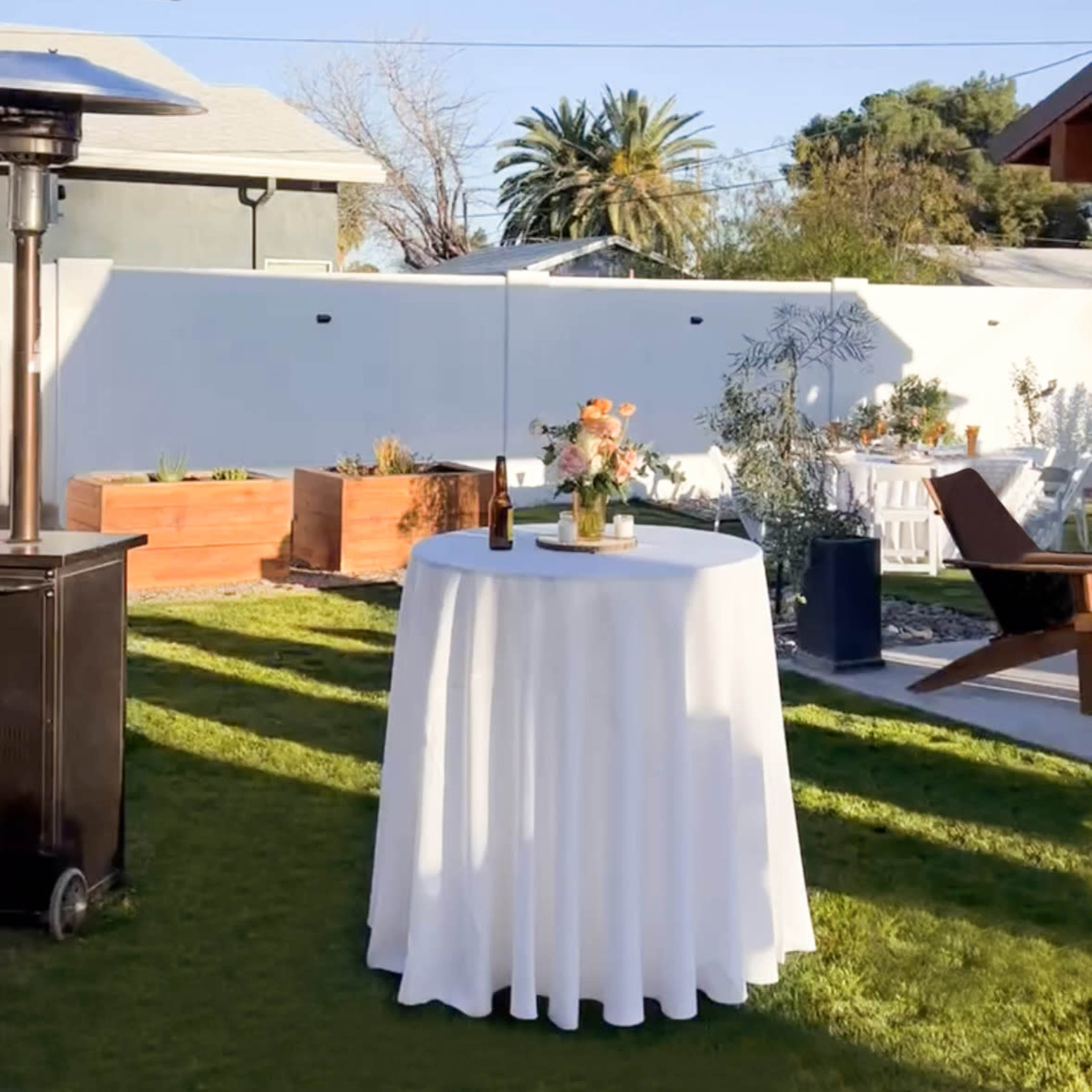 A circular table with a white tablecloth is set up in a grassy backyard, surrounded by planters and a patio heater, under a clear blue sky.