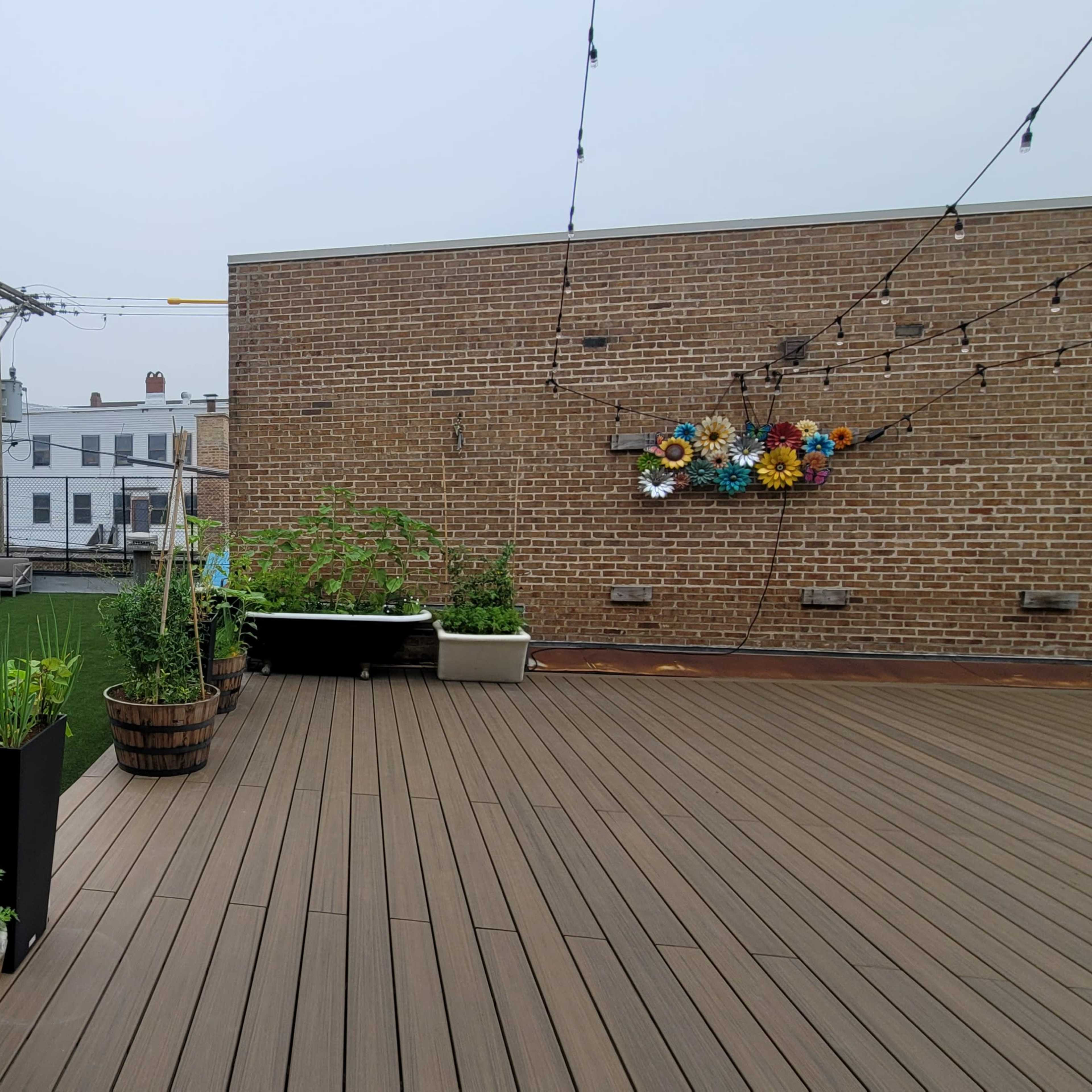 A rooftop garden with wooden decking, planters filled with plants, and a view of brick walls and buildings in the background.