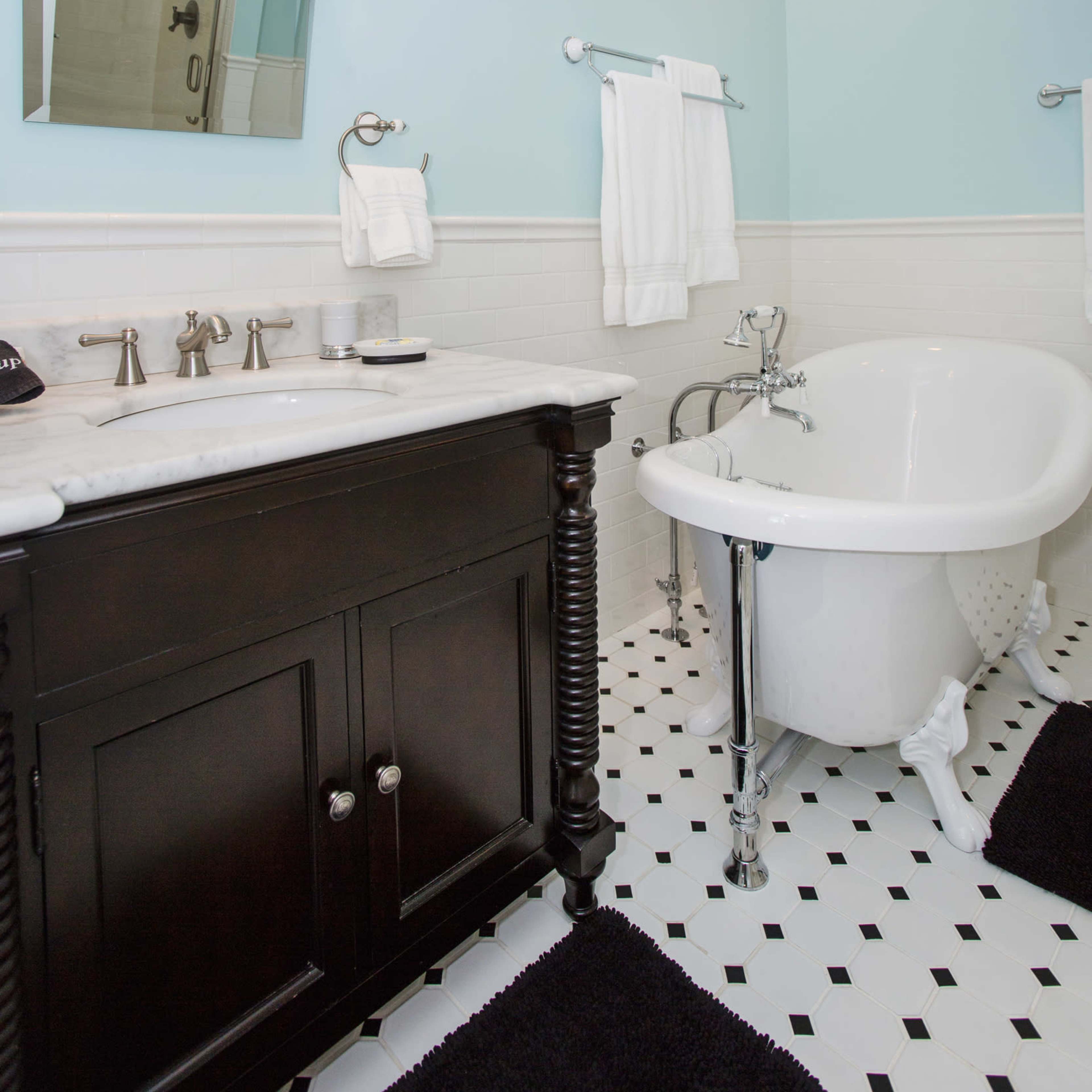 A light blue bathroom with a white clawfoot tub, a dark wooden vanity, and black and white tiled flooring.