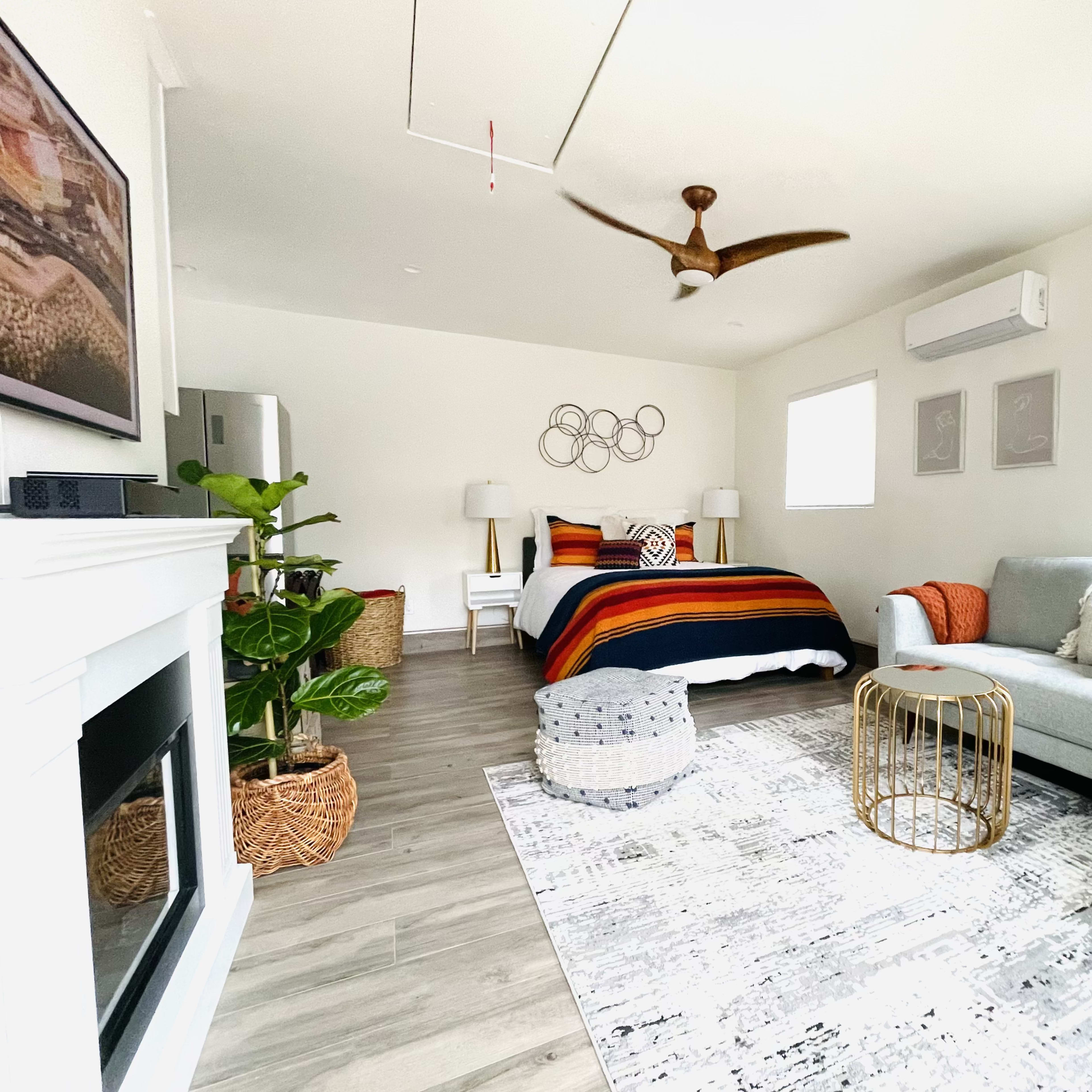 A neatly arranged bedroom with a bed covered in a striped blanket, a green plant in the corner, a light gray sofa, and decorative pillows, set against light-colored walls and wooden flooring.