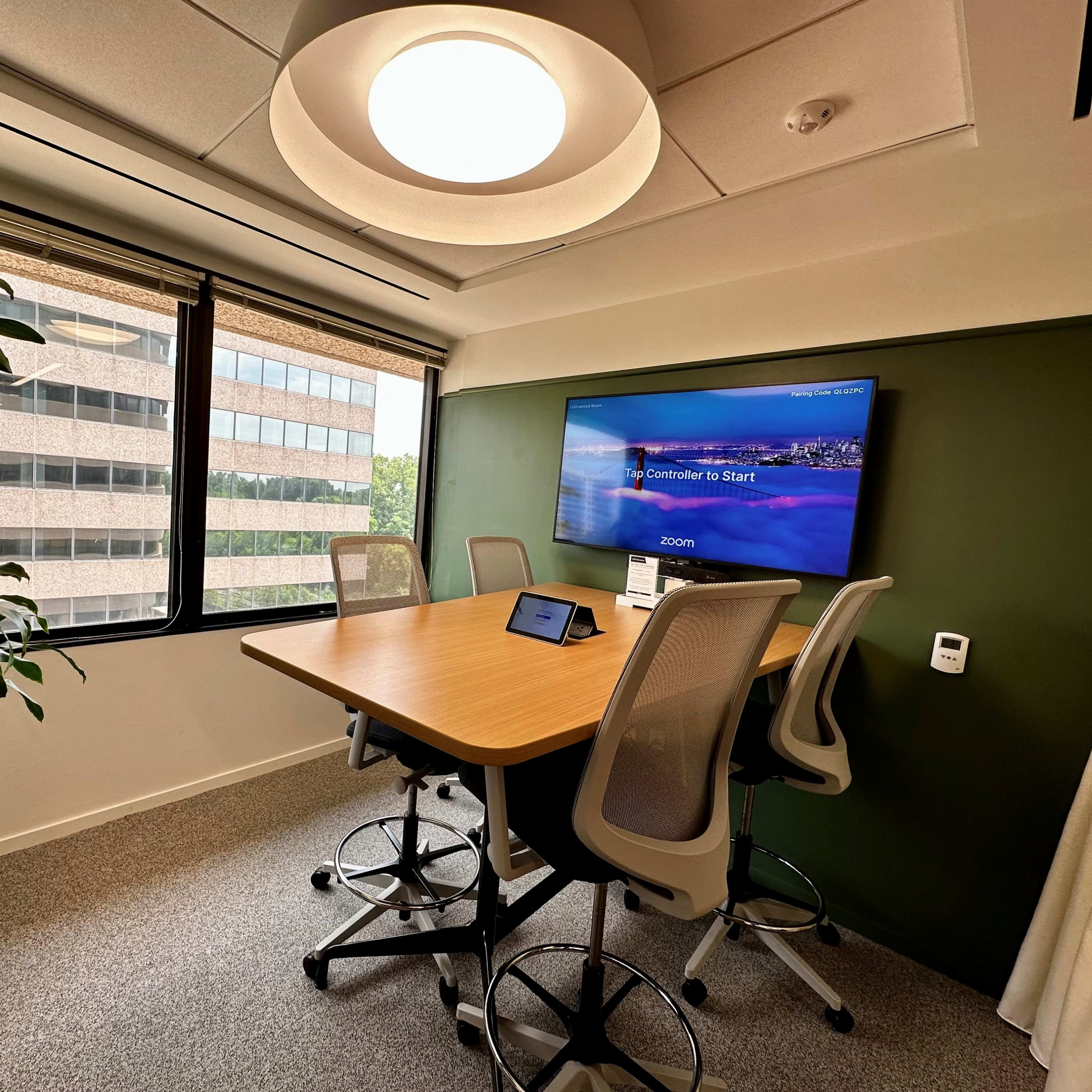A modern conference room features a large table with high-backed chairs, a digital screen displaying a Zoom interface, and large windows providing natural light.