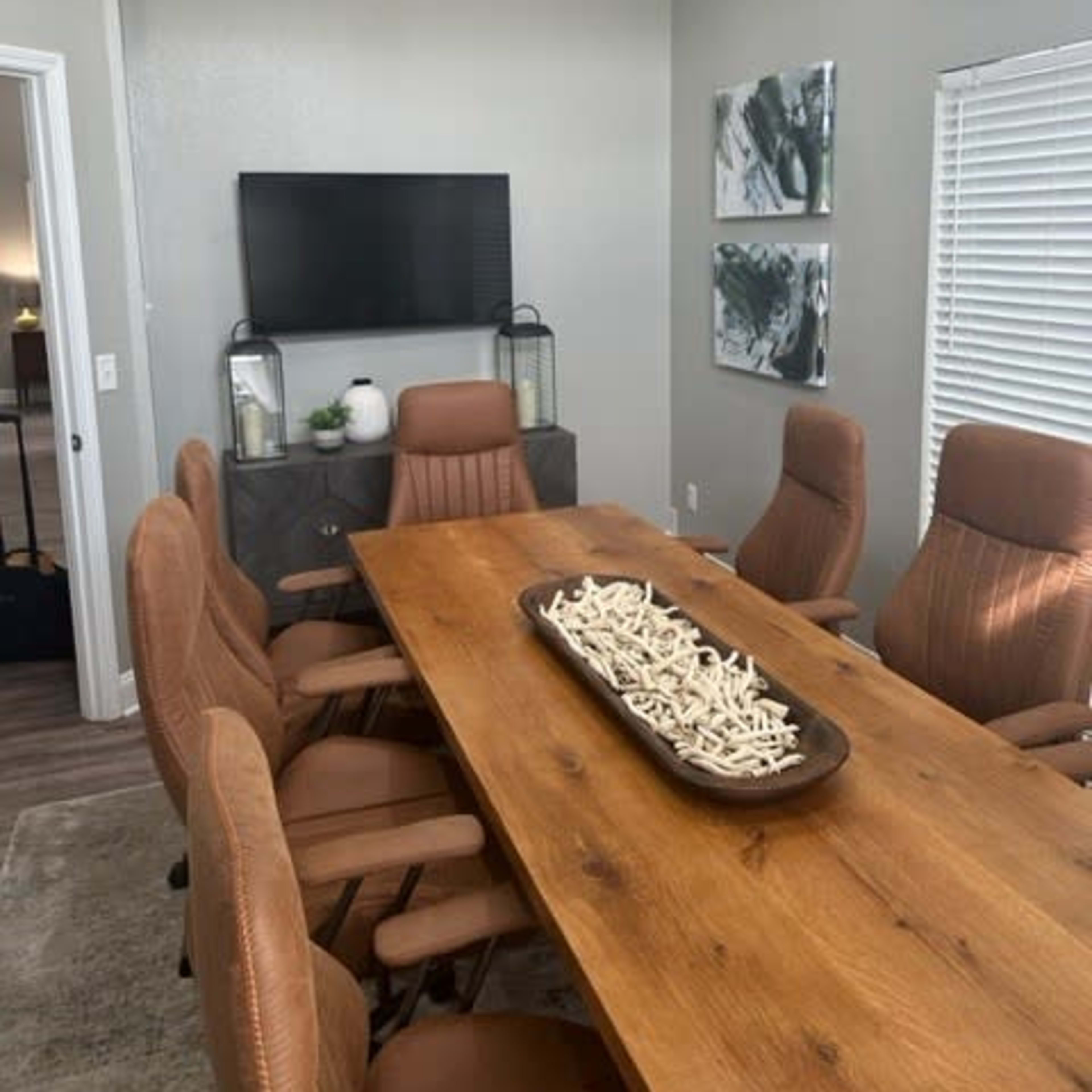 A modern conference room features a large wooden table surrounded by brown leather chairs, with a television mounted on the wall and decorative art on display.
