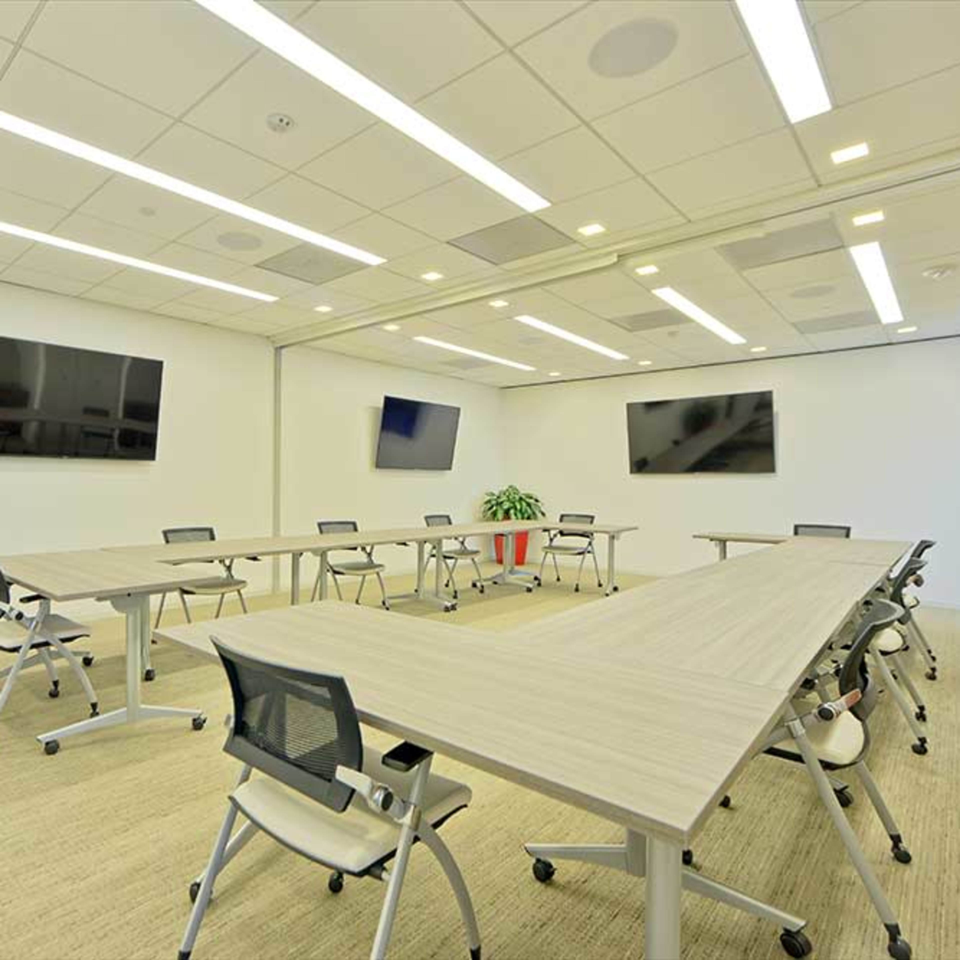 A modern conference room with multiple tables arranged in a U-shape, several televisions mounted on the walls, and a large potted plant in the corner.