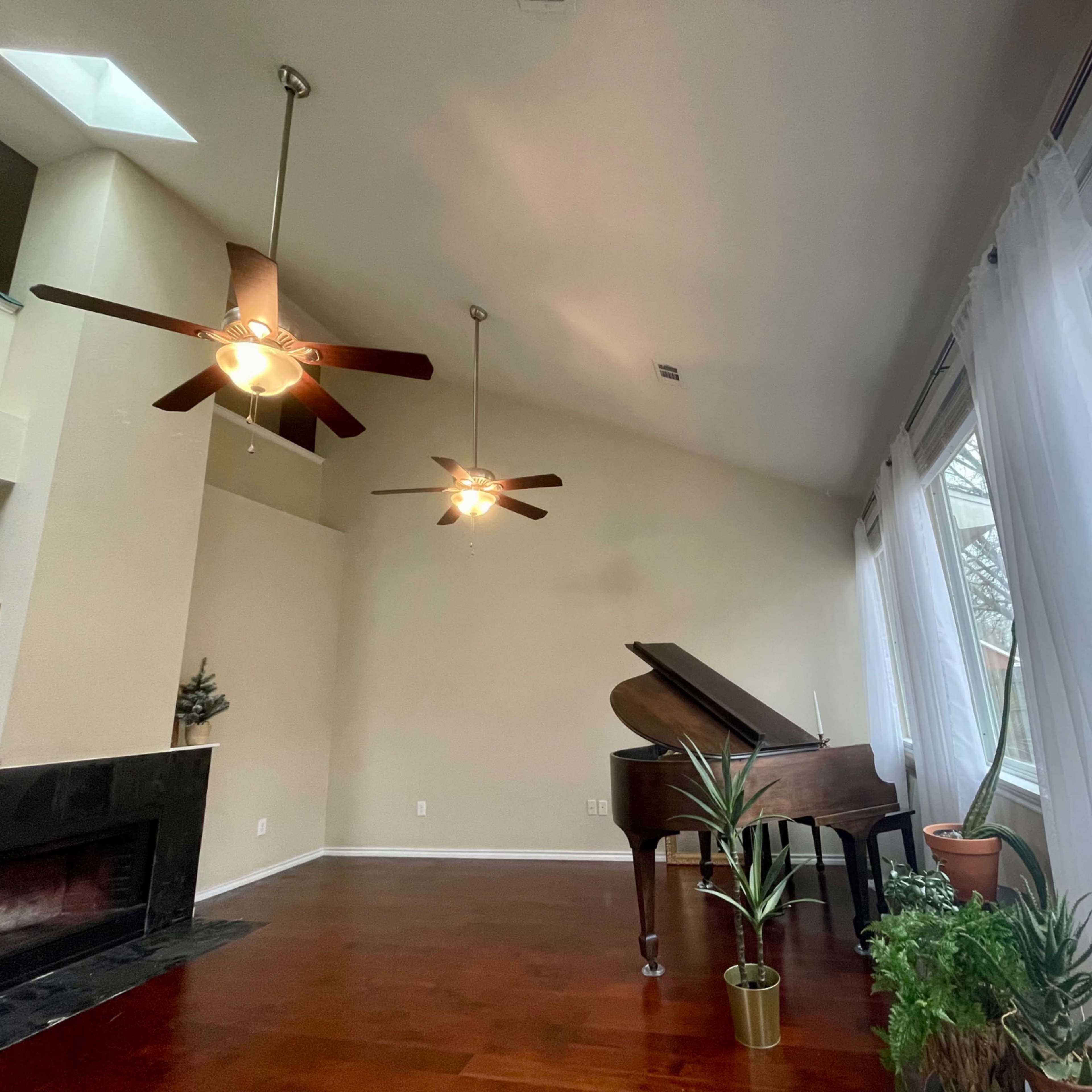 A spacious room features a grand piano, two ceiling fans, and large windows with sheer curtains overlooking a garden.