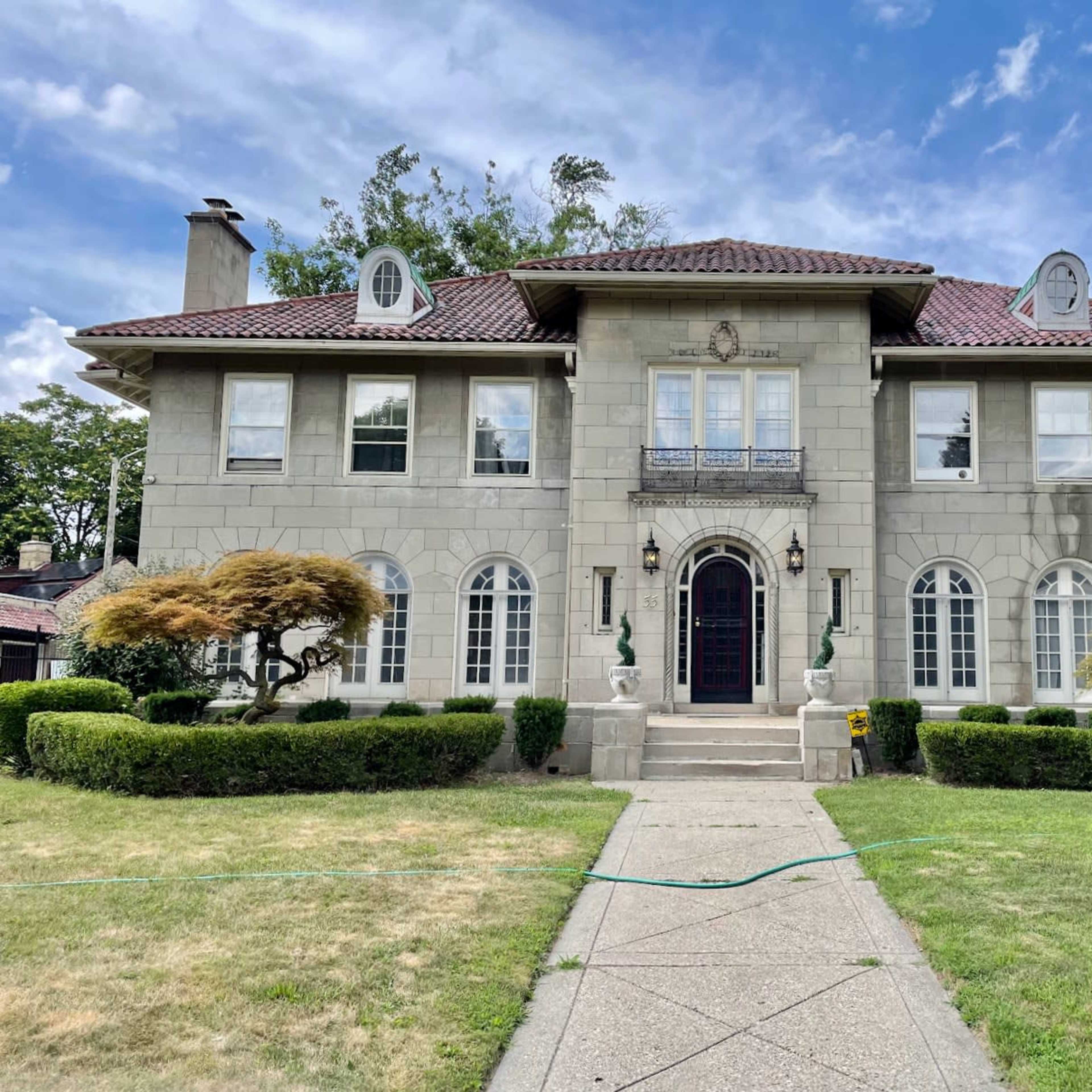 A large, two-story stone house with a tiled roof, flanked by manicured shrubs and two ornamental trees in the front yard.