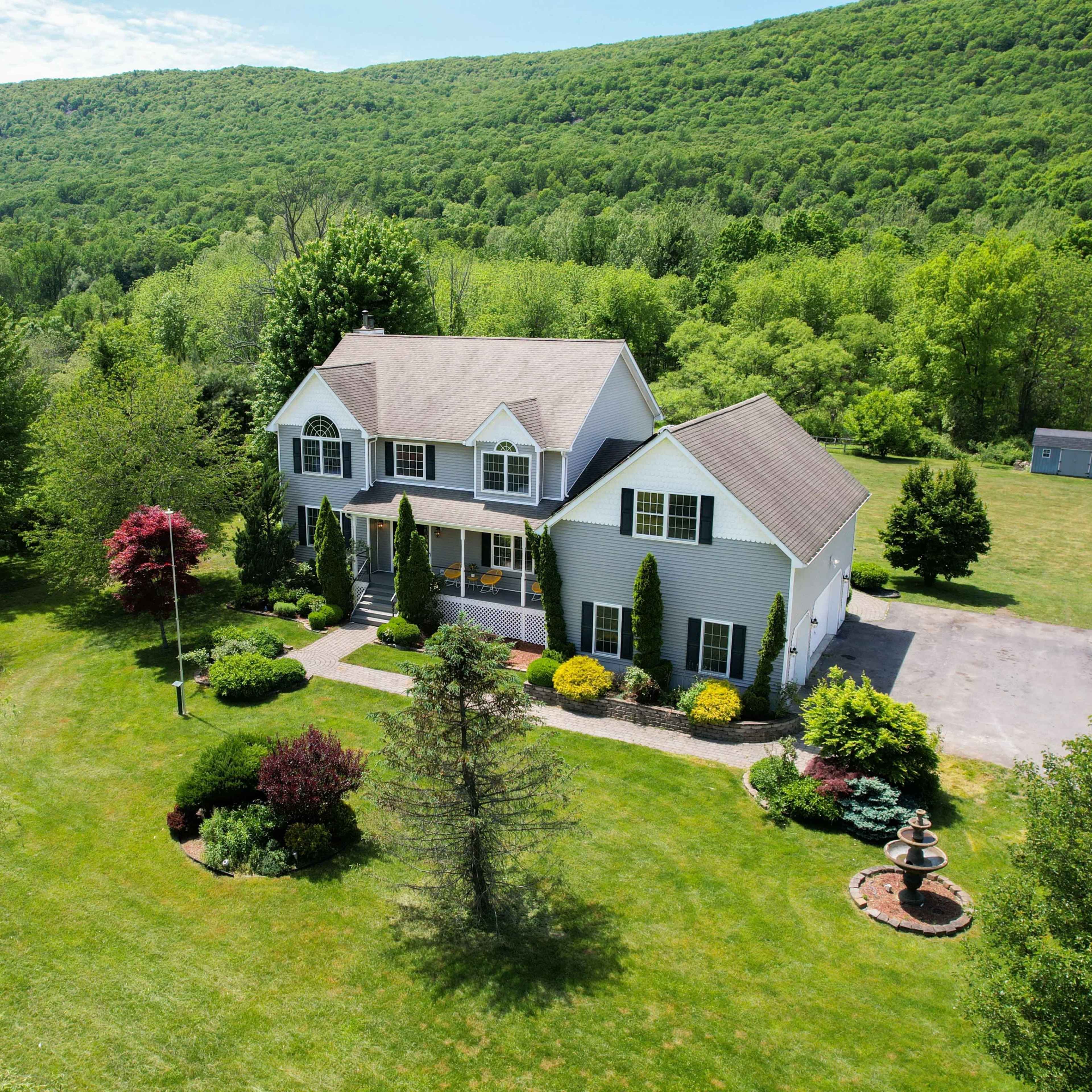 A large two-story house surrounded by landscaped gardens and trees, set against a backdrop of rolling green hills.