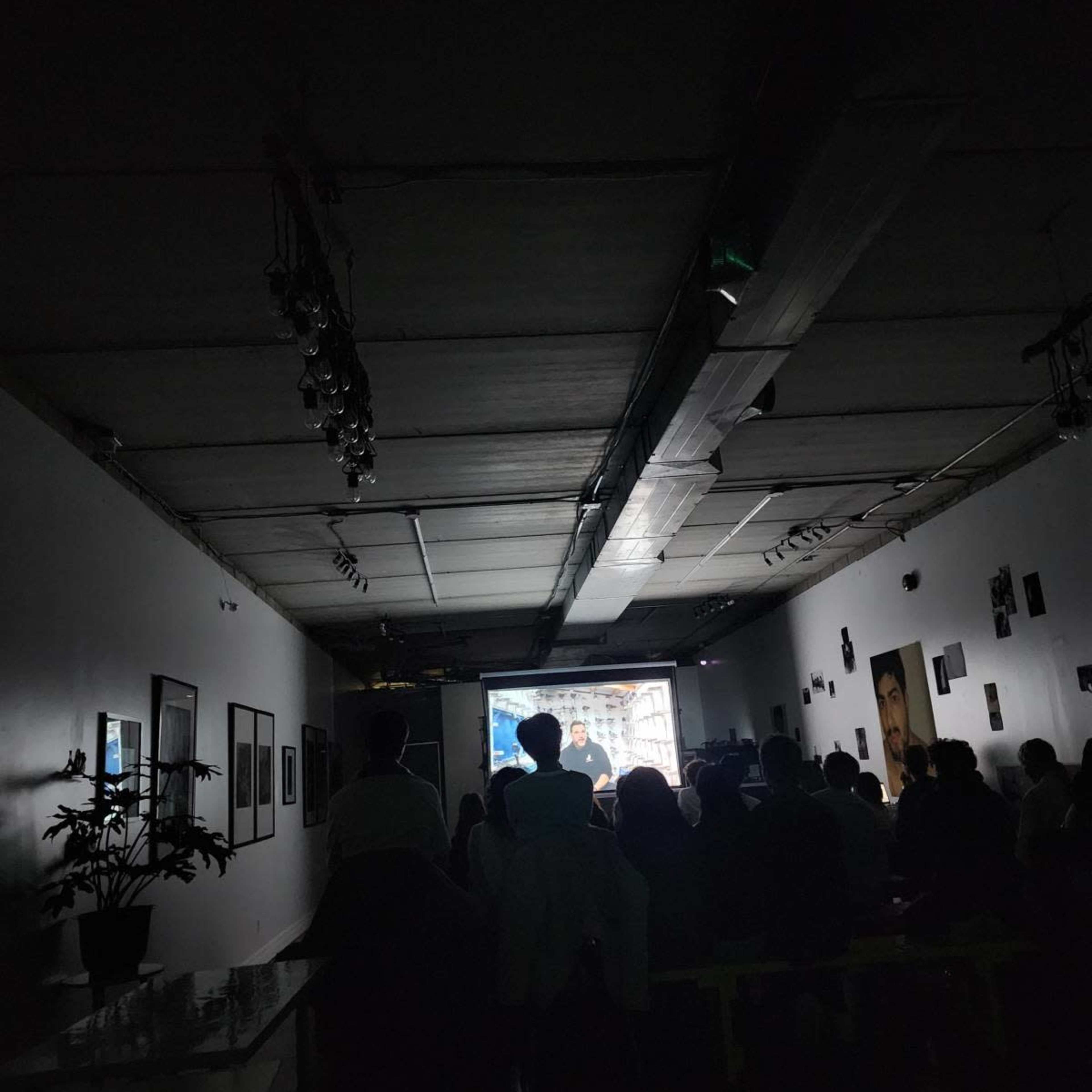 A group of people is seated in a dimly lit room watching a presentation projected on a screen.