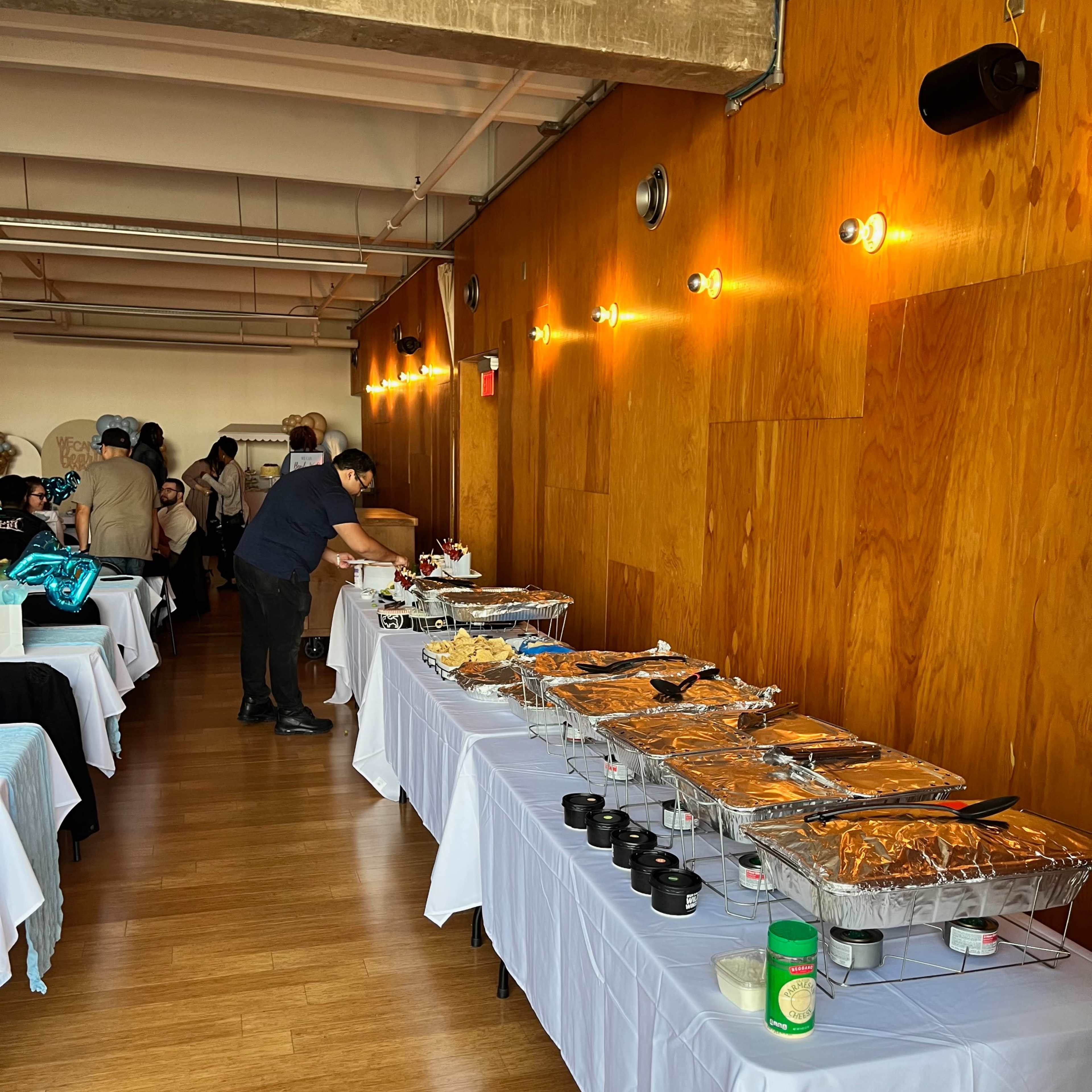 A buffet display with multiple tables set up along a wall, featuring trays of food and guests seated in the background.