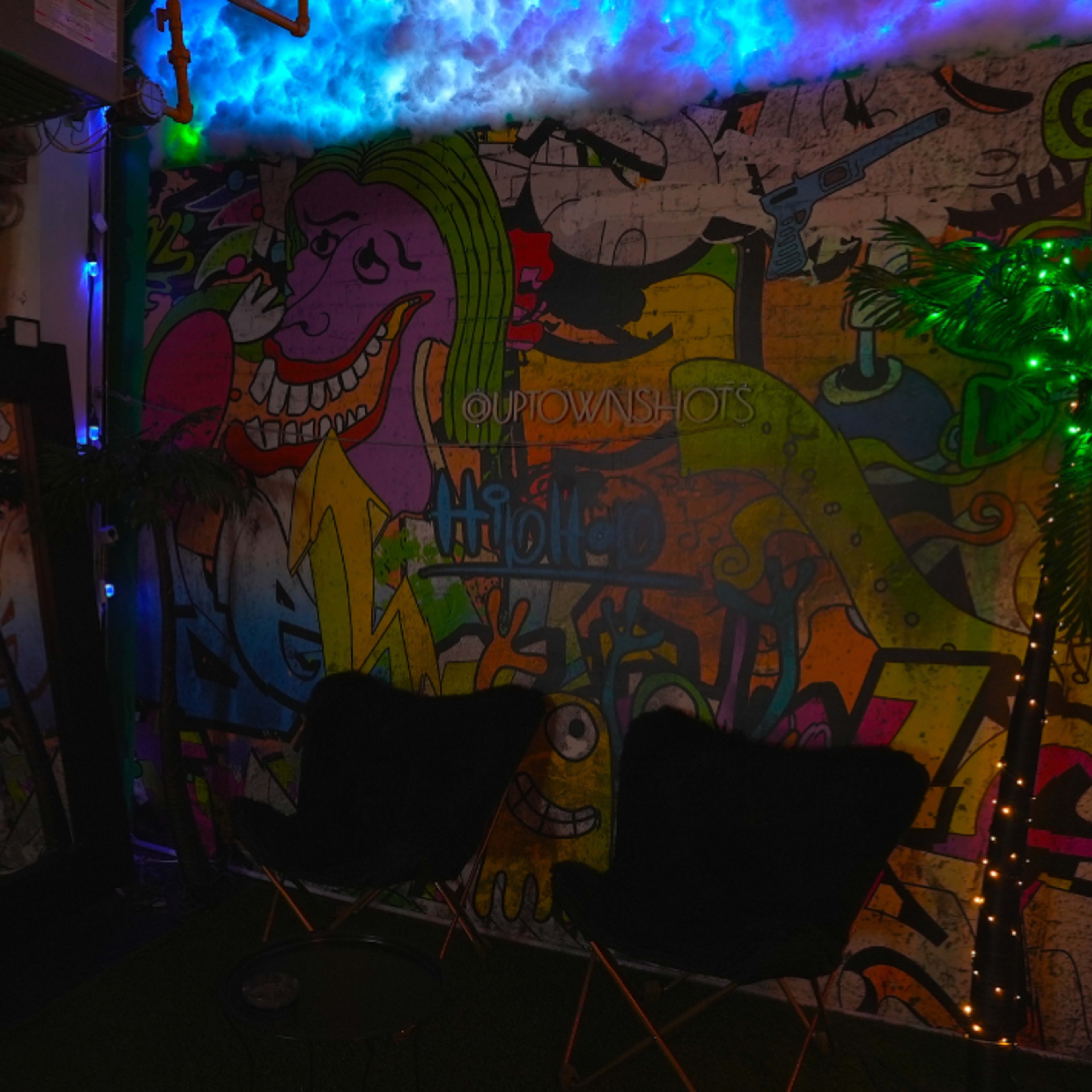 A dimly lit room with a colorful graffiti wall, two black chairs, a mirror, and a small illuminated palm tree.