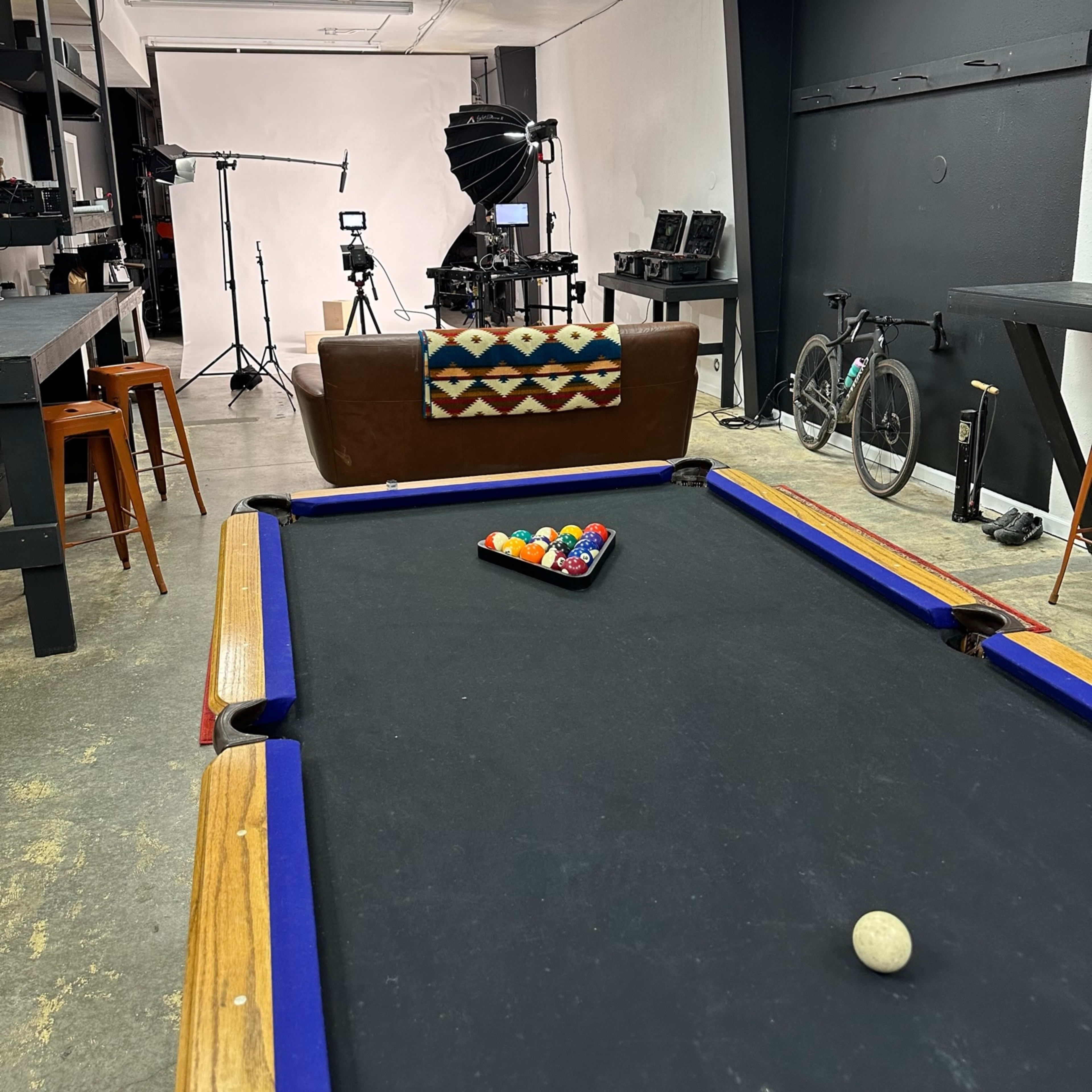 A room with a pool table in the foreground, a brown couch in the center, and various photography equipment and a bicycle in the background.