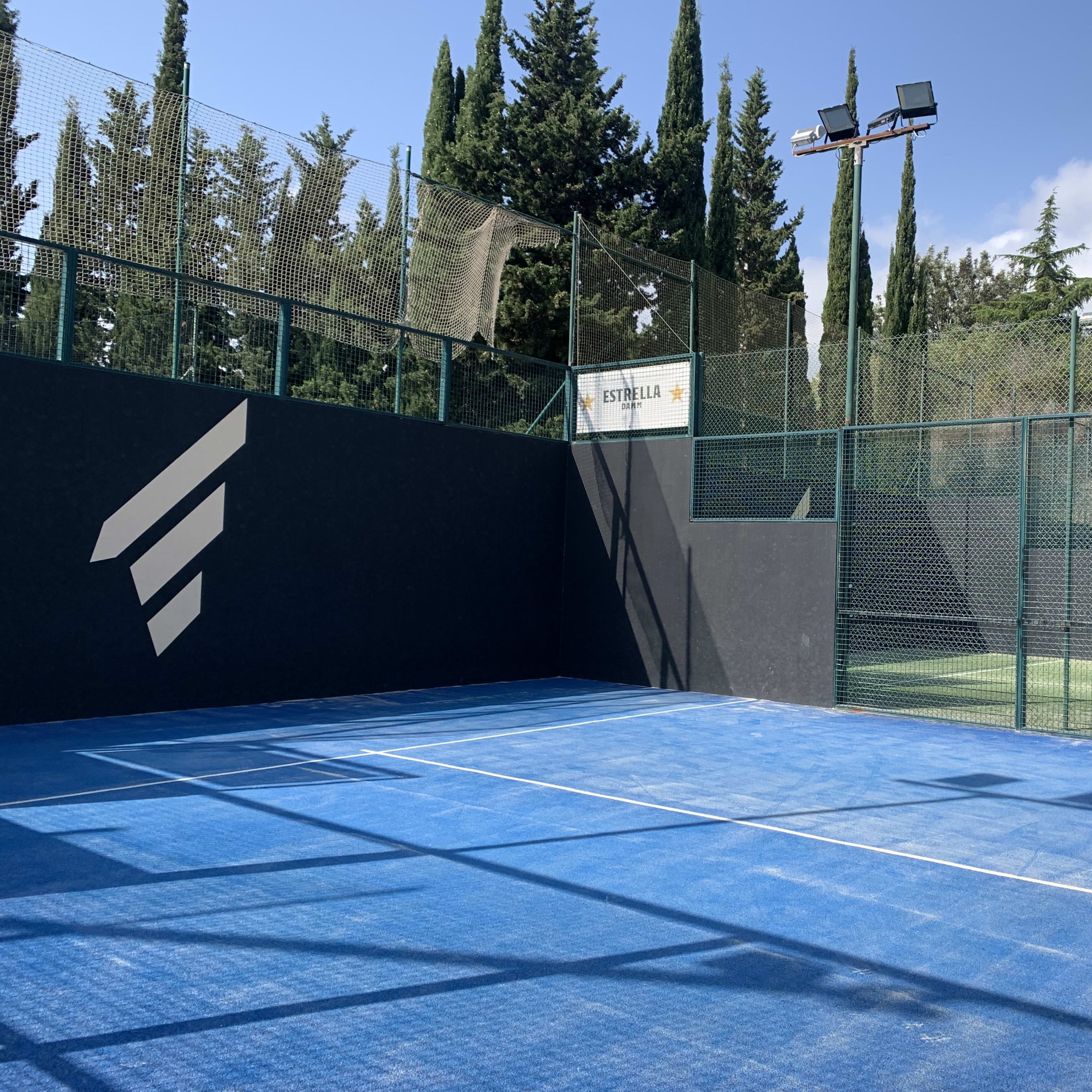 A blue padel court enclosed by a green mesh fence and surrounded by tall trees under a partly cloudy sky.