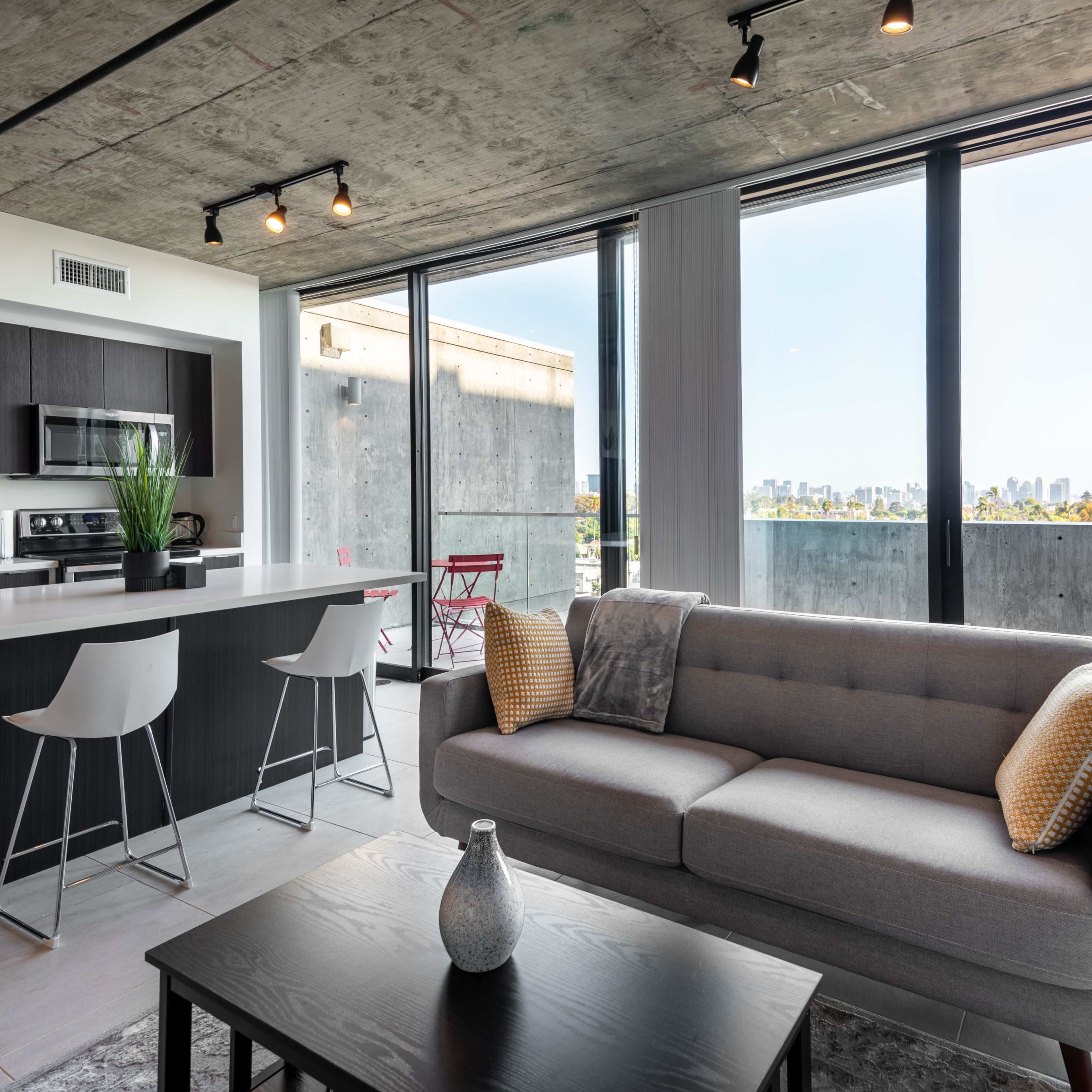 A modern apartment features a kitchen and living area with a view of the city skyline through large windows.