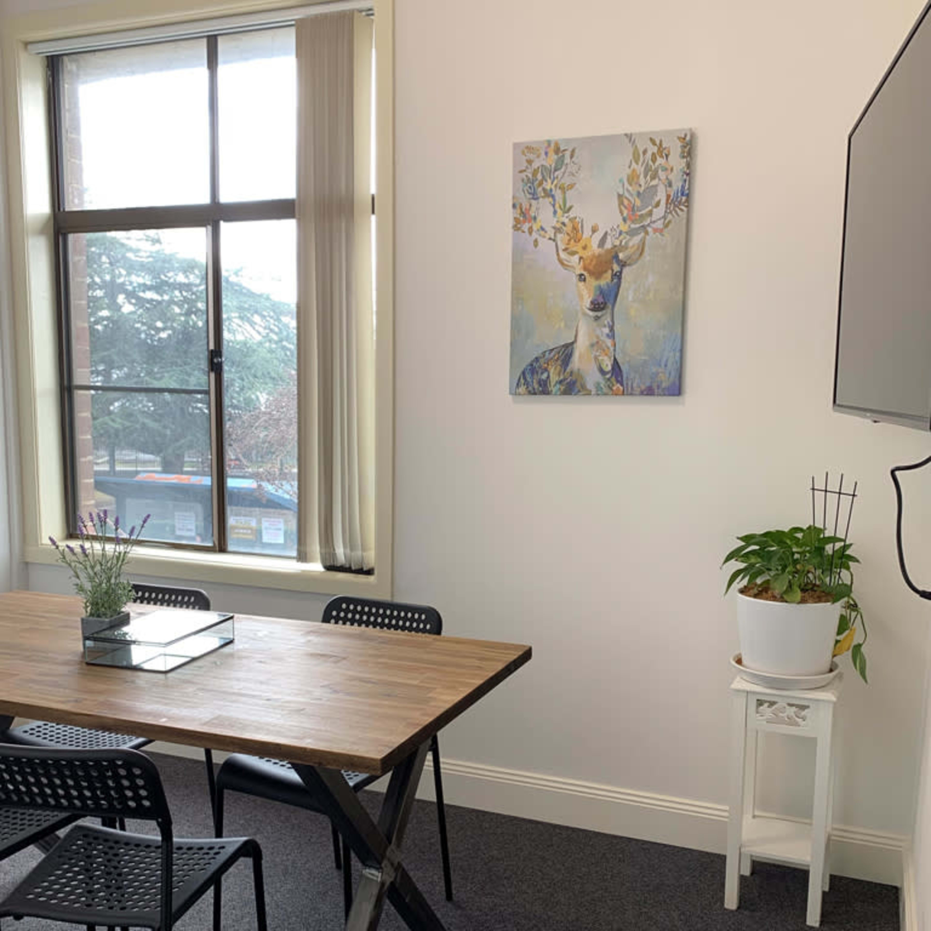 A small meeting room with a wooden table, black chairs, a deer painting on the wall, and a plant on a stand near a window.