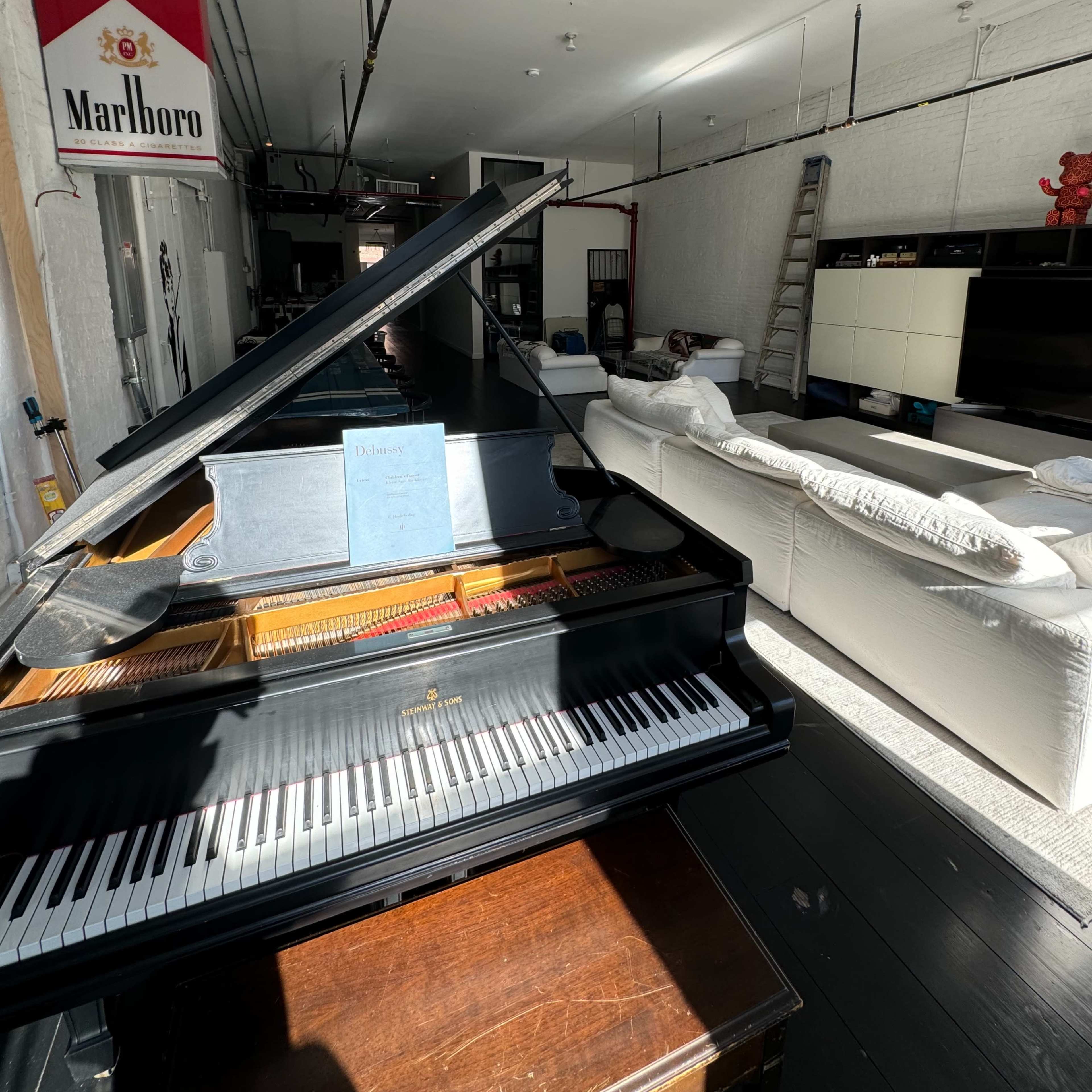 A grand piano in the foreground with a living space that includes white couches and a TV in the background, illuminated by natural light.