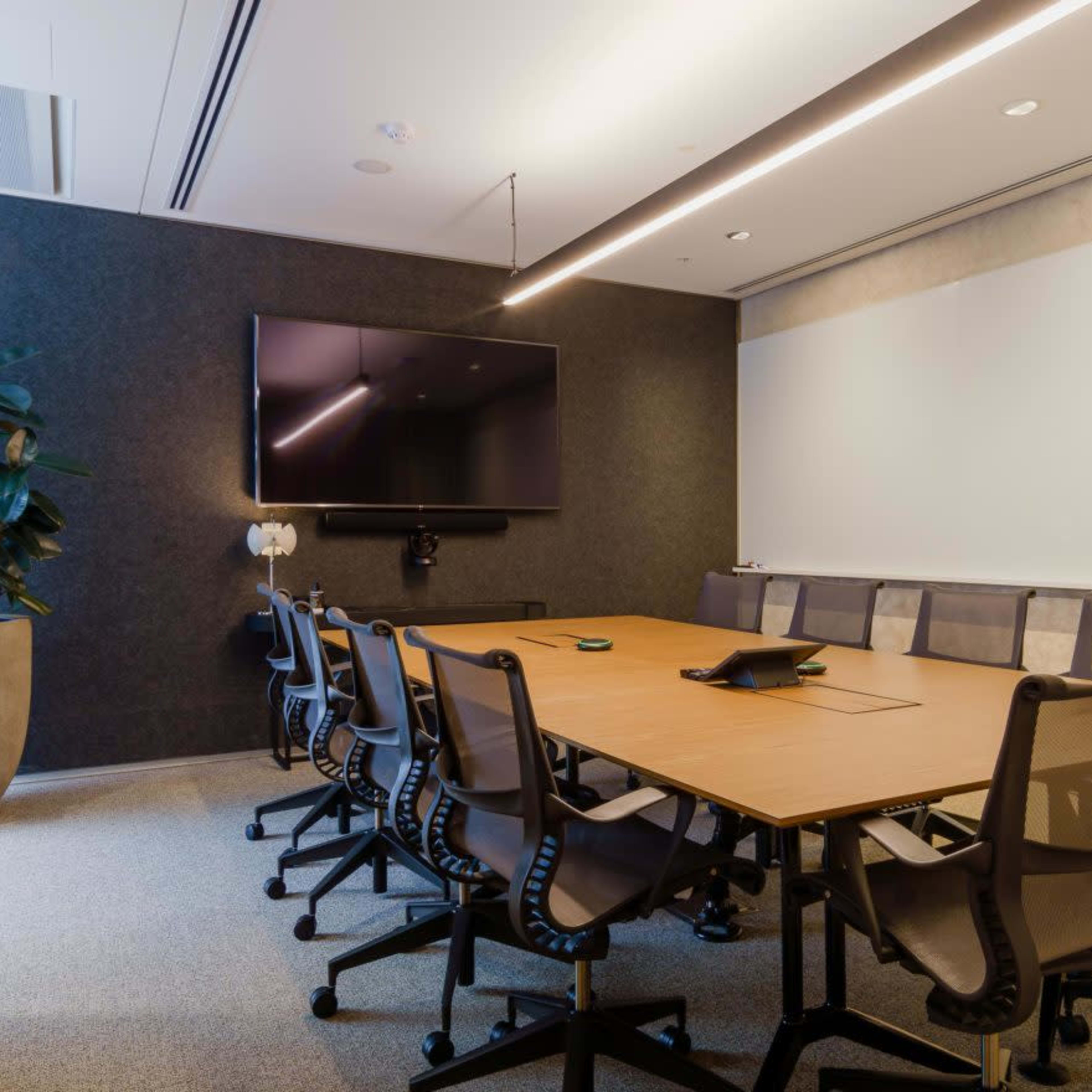 A modern conference room features a large table surrounded by ergonomic chairs, a television screen mounted on the wall, and a potted plant in the corner.