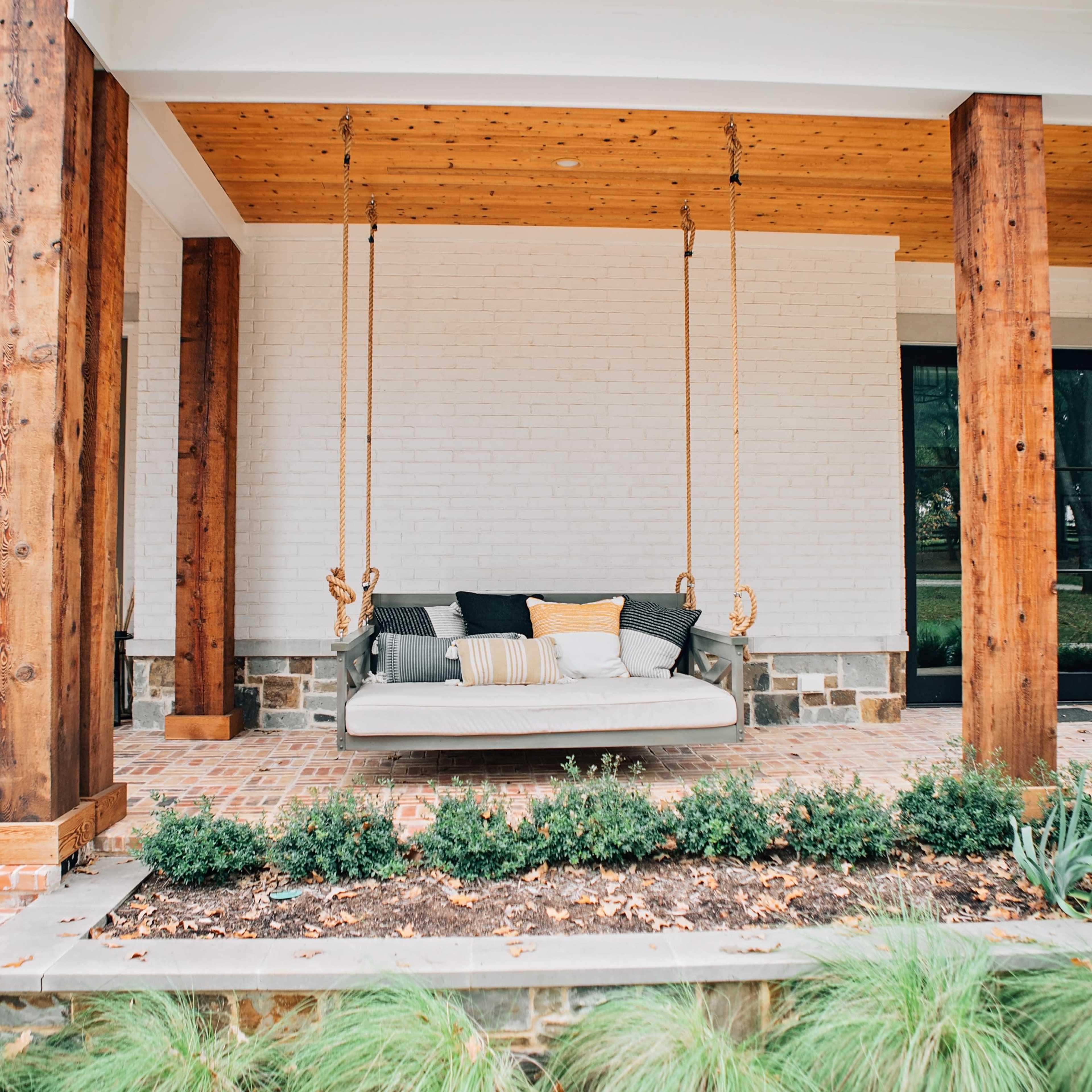 A porch swing is suspended between wooden posts on a covered patio, with a brick floor and greenery surrounding it.