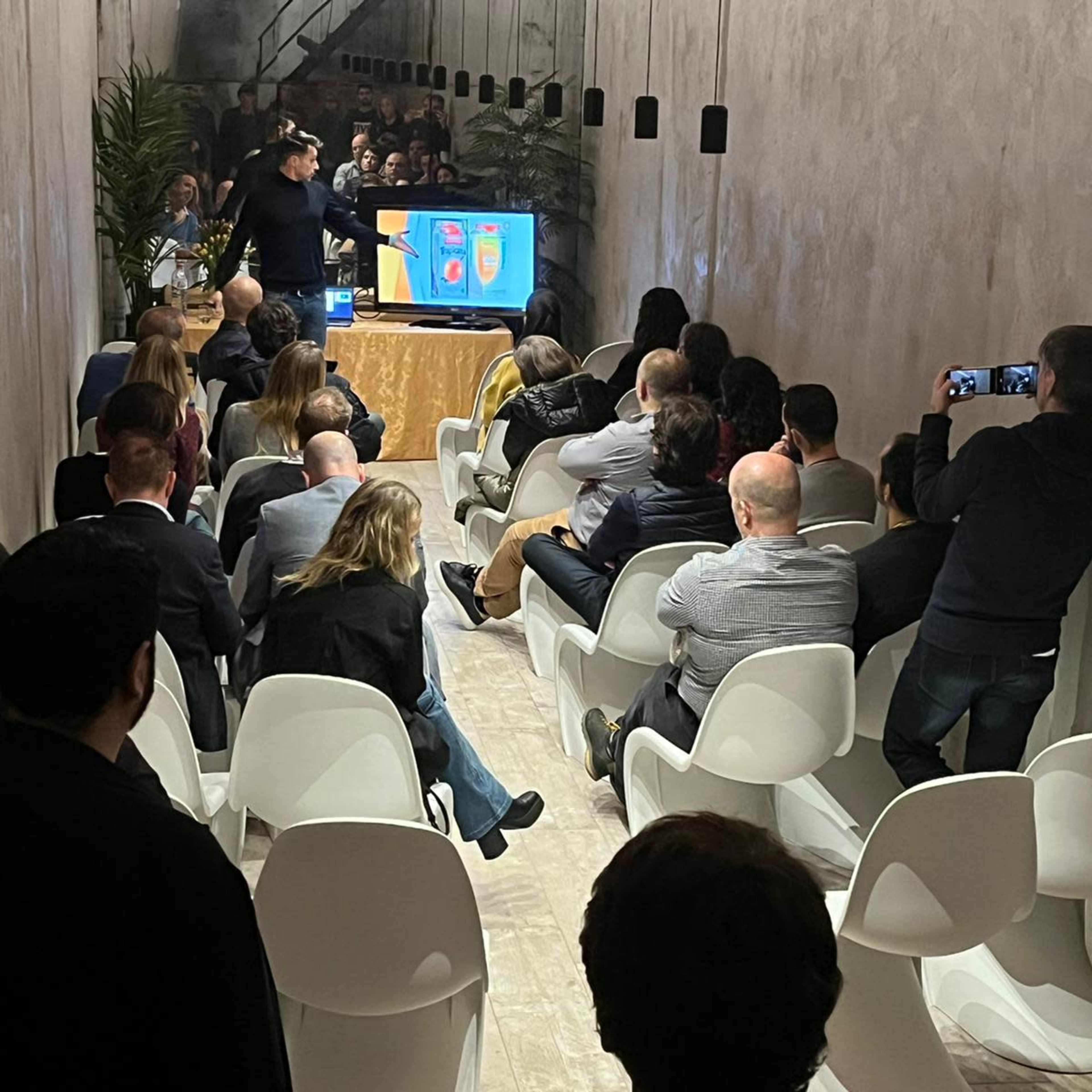 A group of people are seated in a narrow room, facing a presenter who stands in front of a screen displaying colorful graphics.