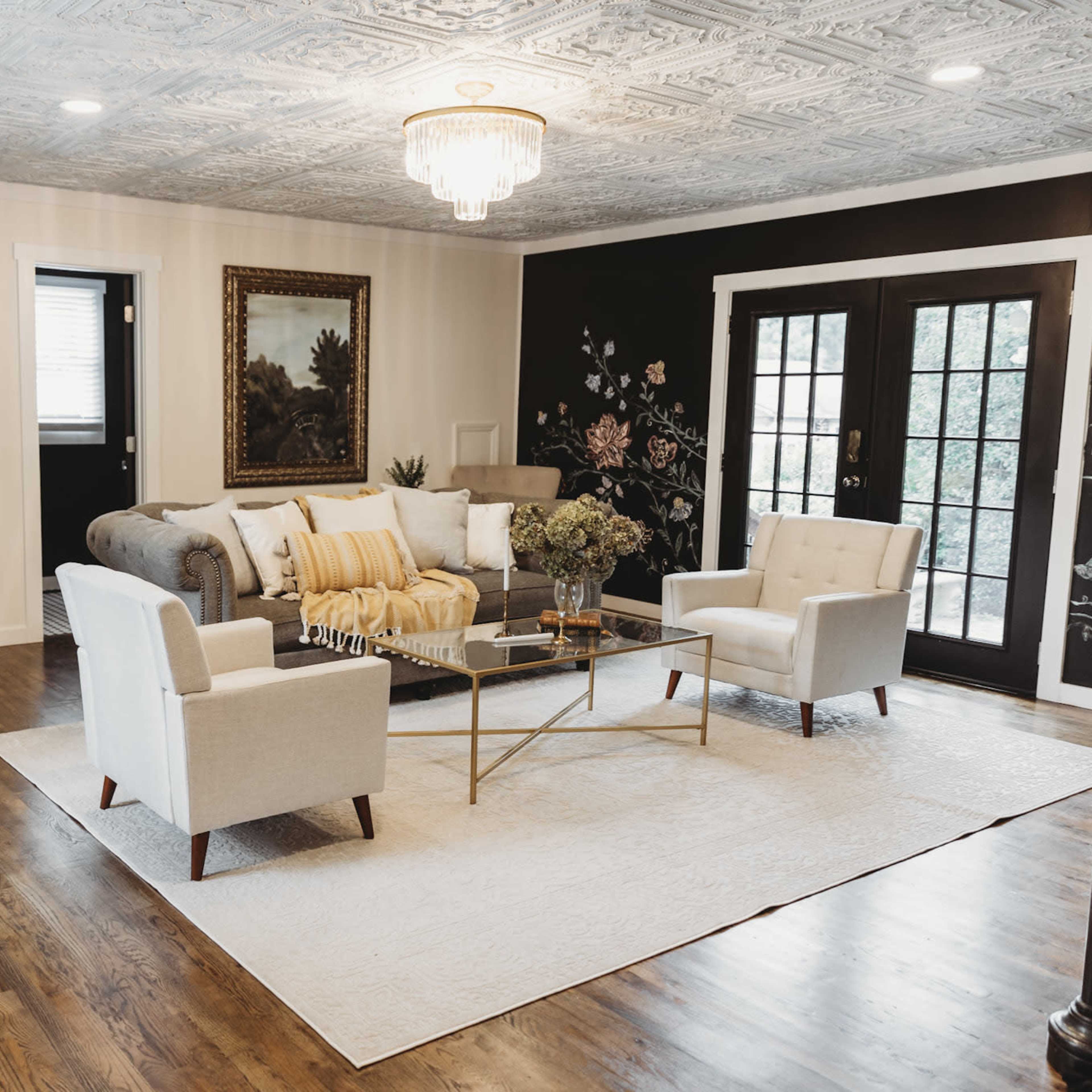 A modern living room with a gray sofa, two white armchairs, a glass coffee table, and large windows leading to an outdoor area.