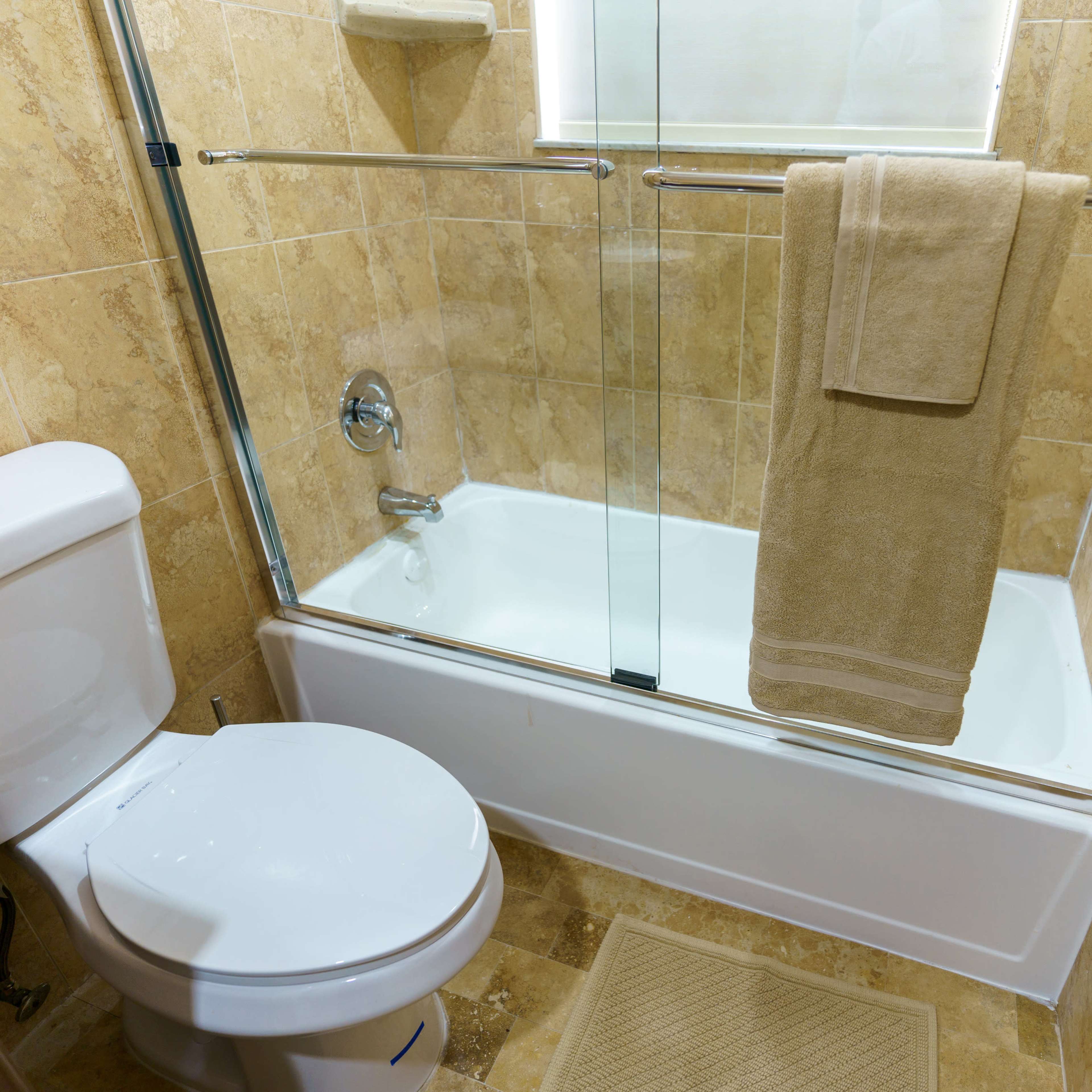 A clean bathroom featuring a glass shower enclosure, a white bathtub, a toilet, and beige towels hanging on a rod.