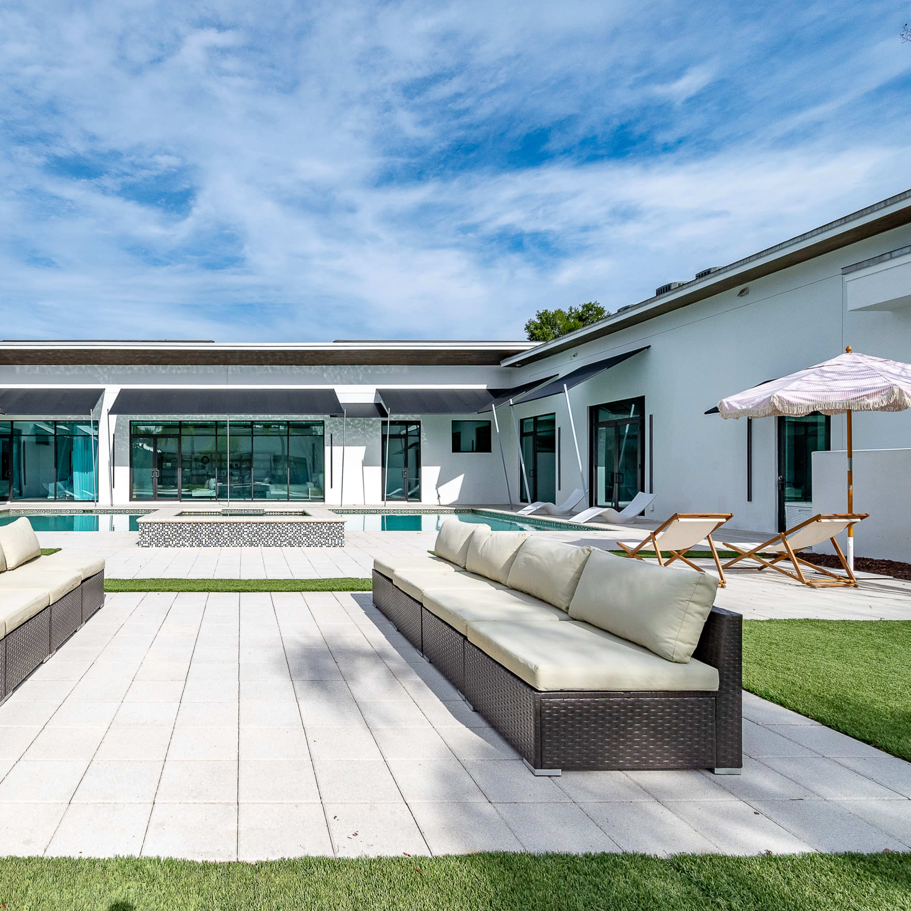 A modern outdoor area with two cream-colored sofas facing a rectangular swimming pool surrounded by a spacious patio and glass walls.