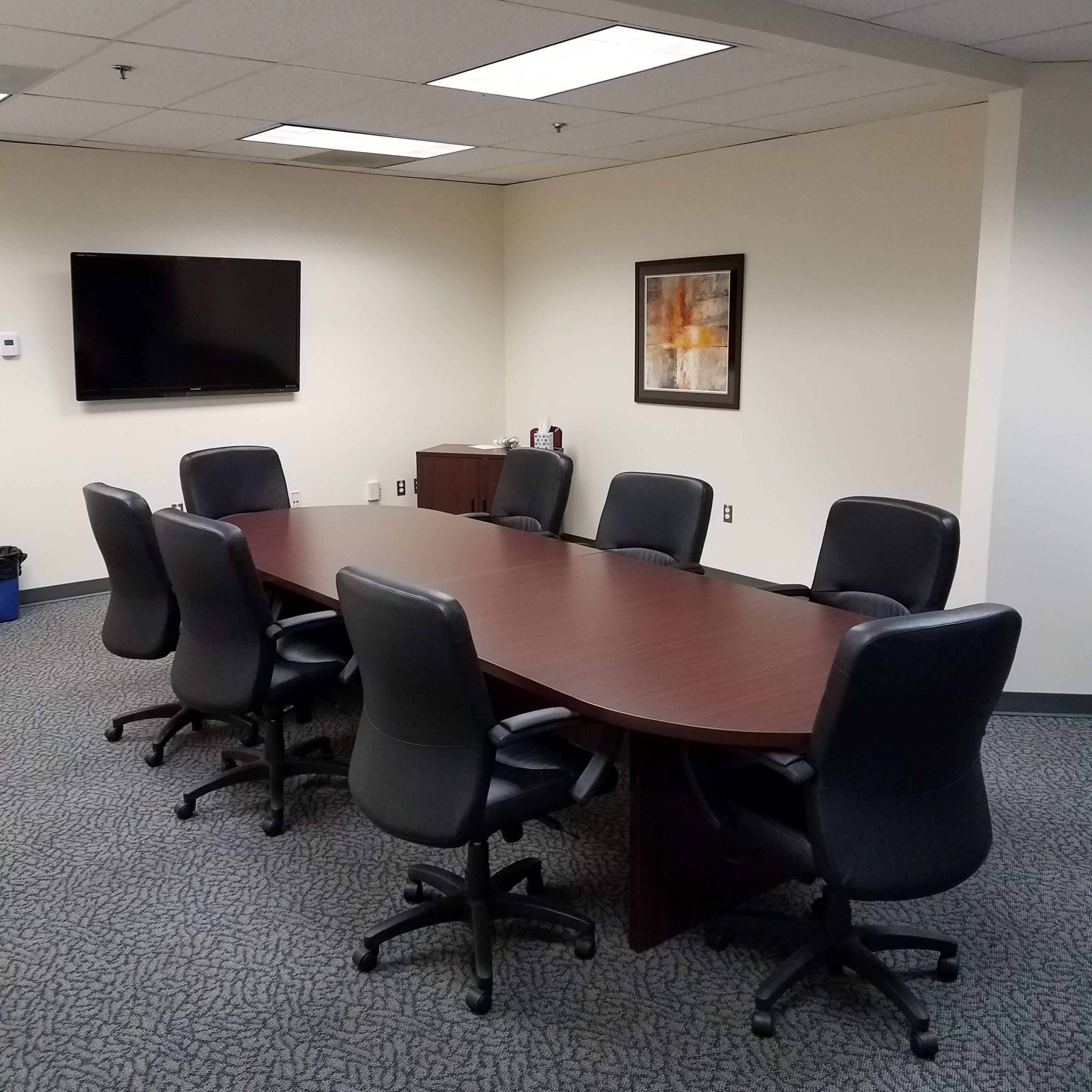 A conference room with a rectangular wooden table surrounded by eight black rolling chairs, a television mounted on the wall, and a piece of framed art.