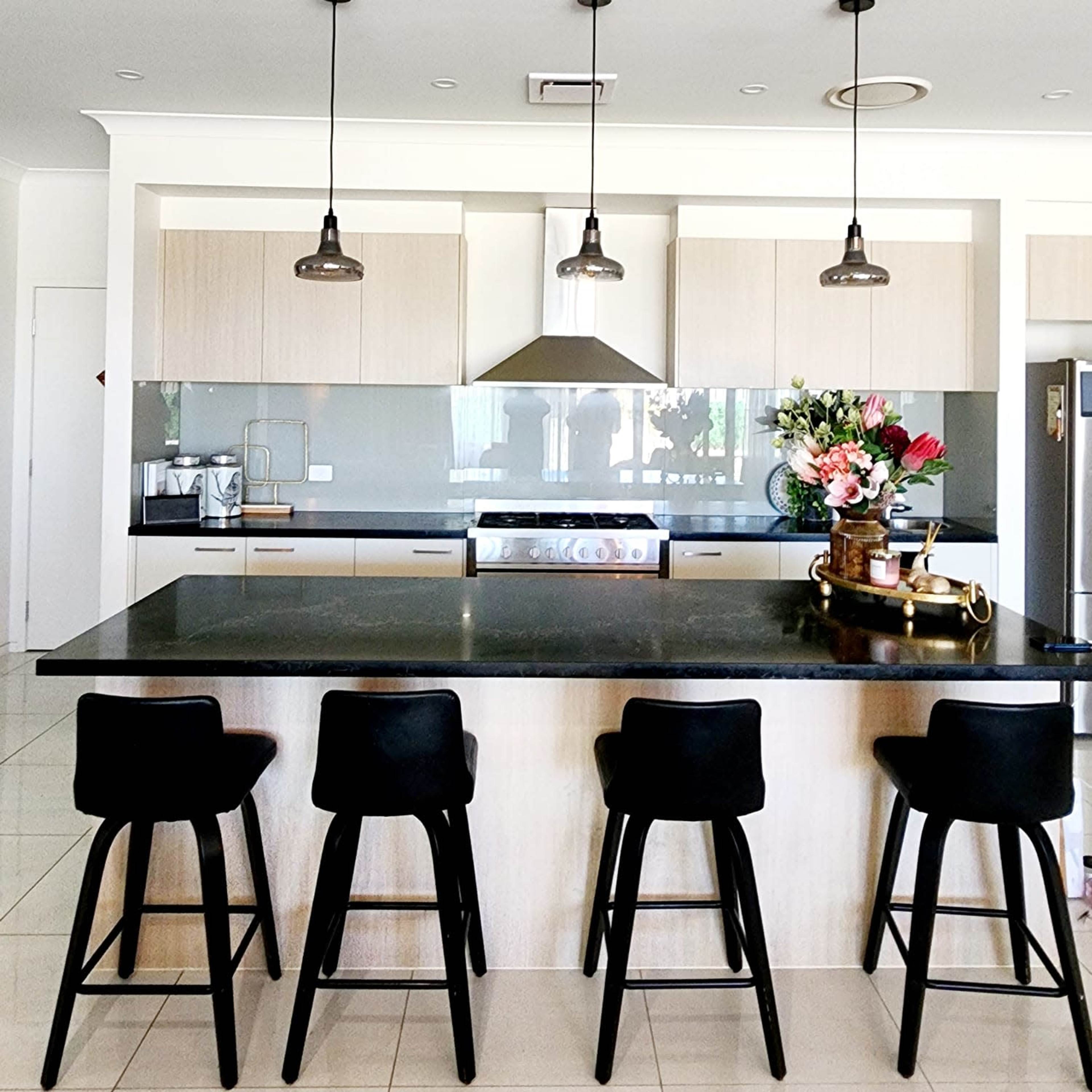 A modern kitchen features a dark countertop and light wooden cabinets, with four black bar stools positioned at a large island and pendant lights hanging above.