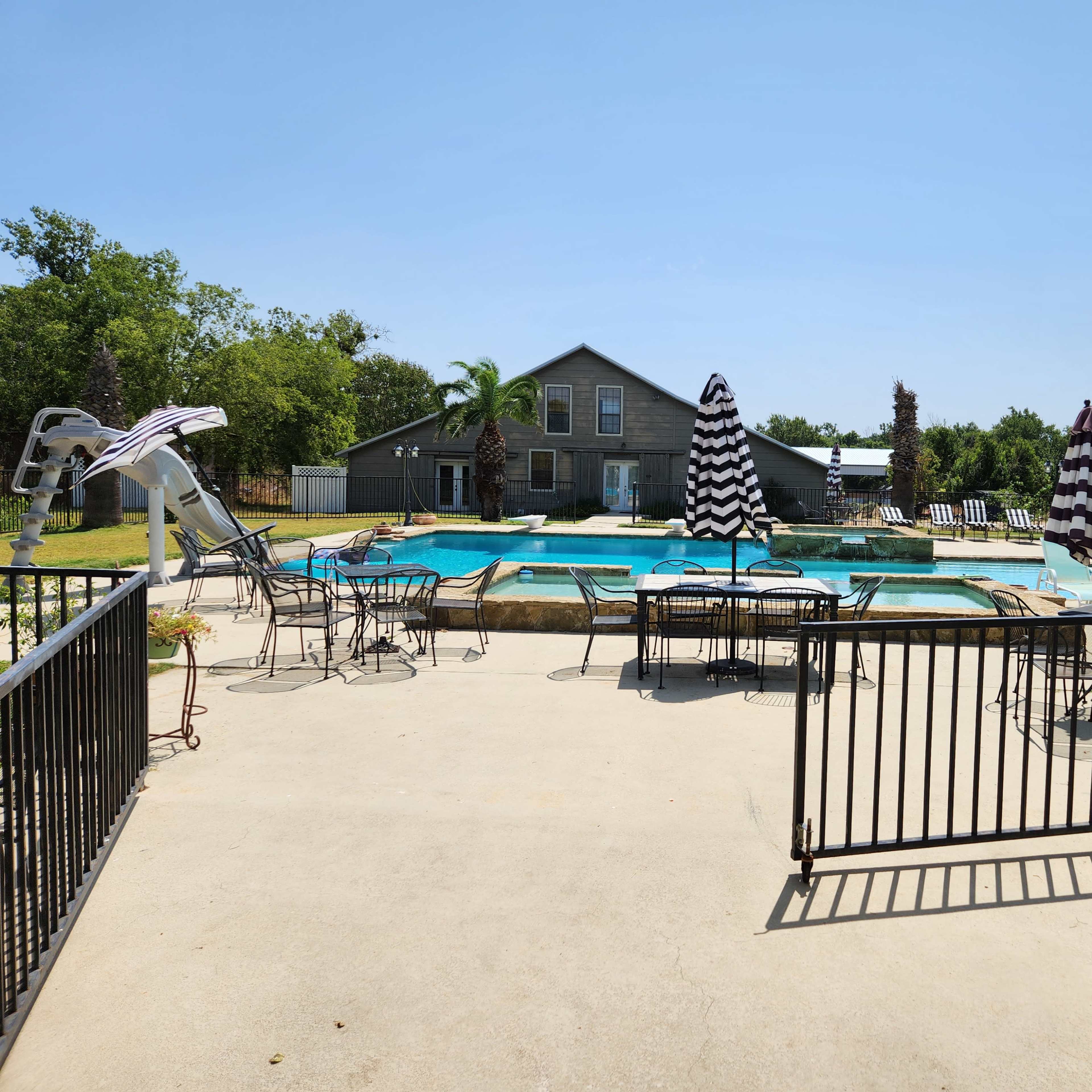 A swimming pool area surrounded by a concrete patio with several chairs and umbrellas, set against a backdrop of greenery and a building.