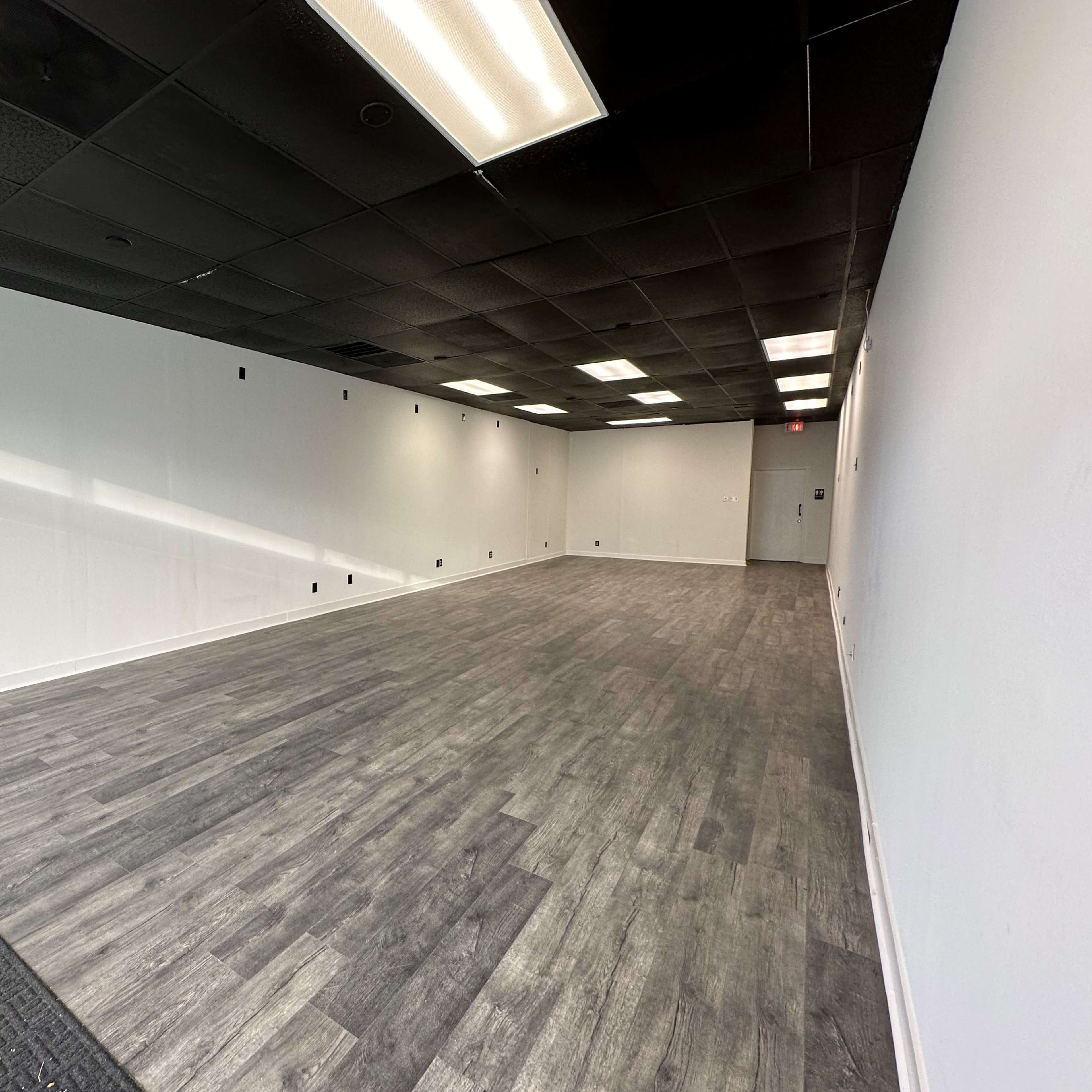A spacious, empty room with gray wooden flooring and white walls, illuminated by overhead lights.