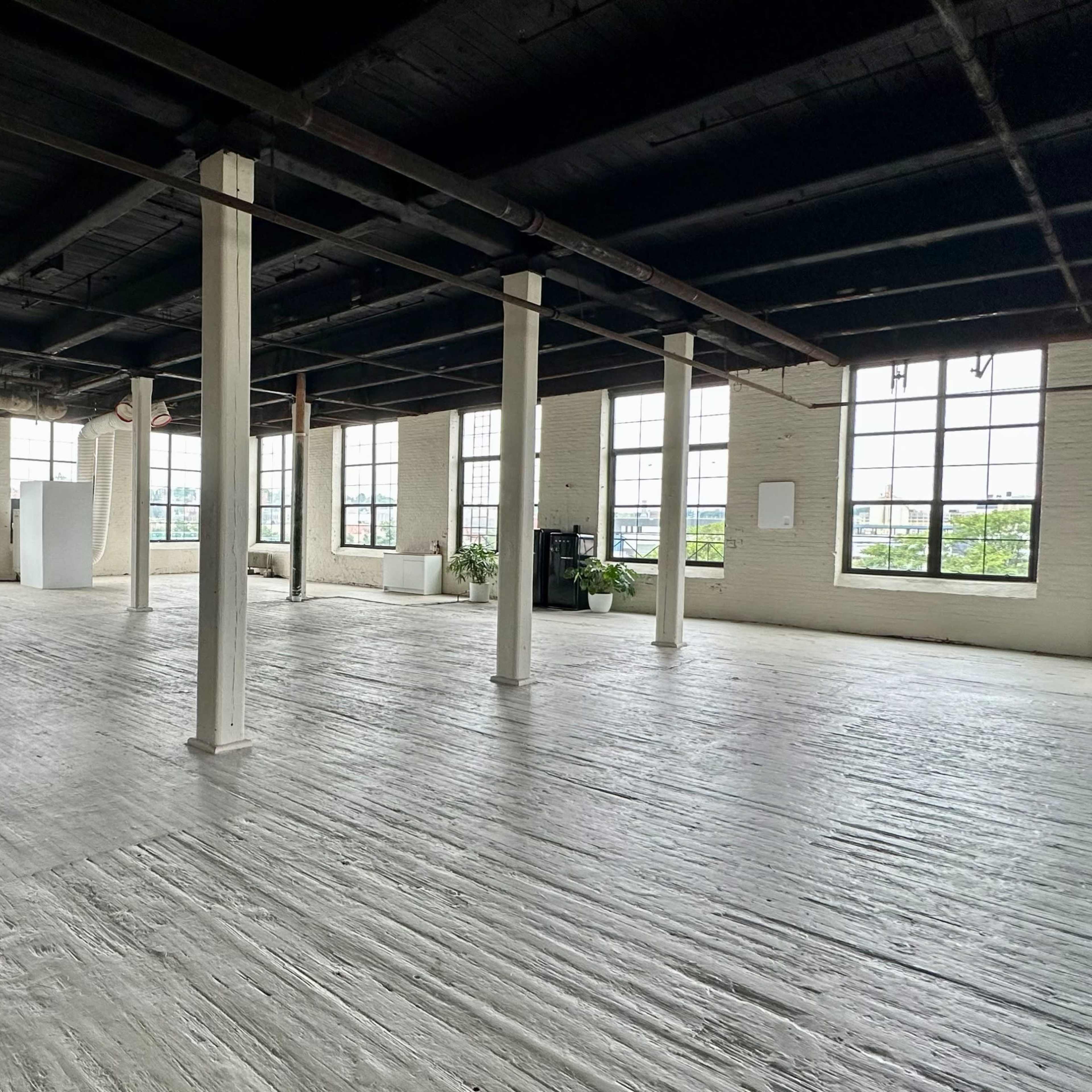 A wide, empty industrial-style room with large windows and wooden flooring.