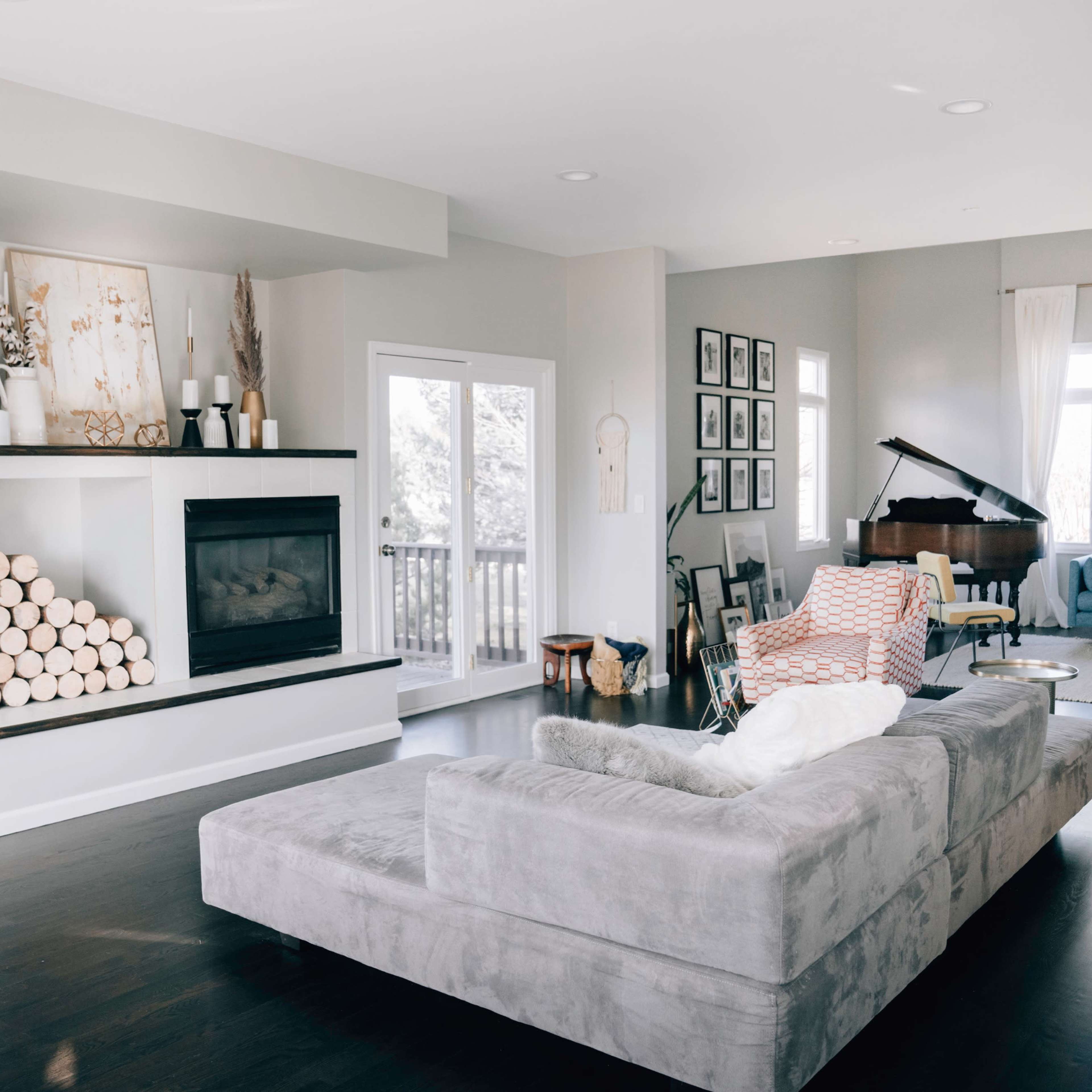 A modern living room with a gray sectional sofa, a fireplace with stacked logs, and a piano near a large window.
