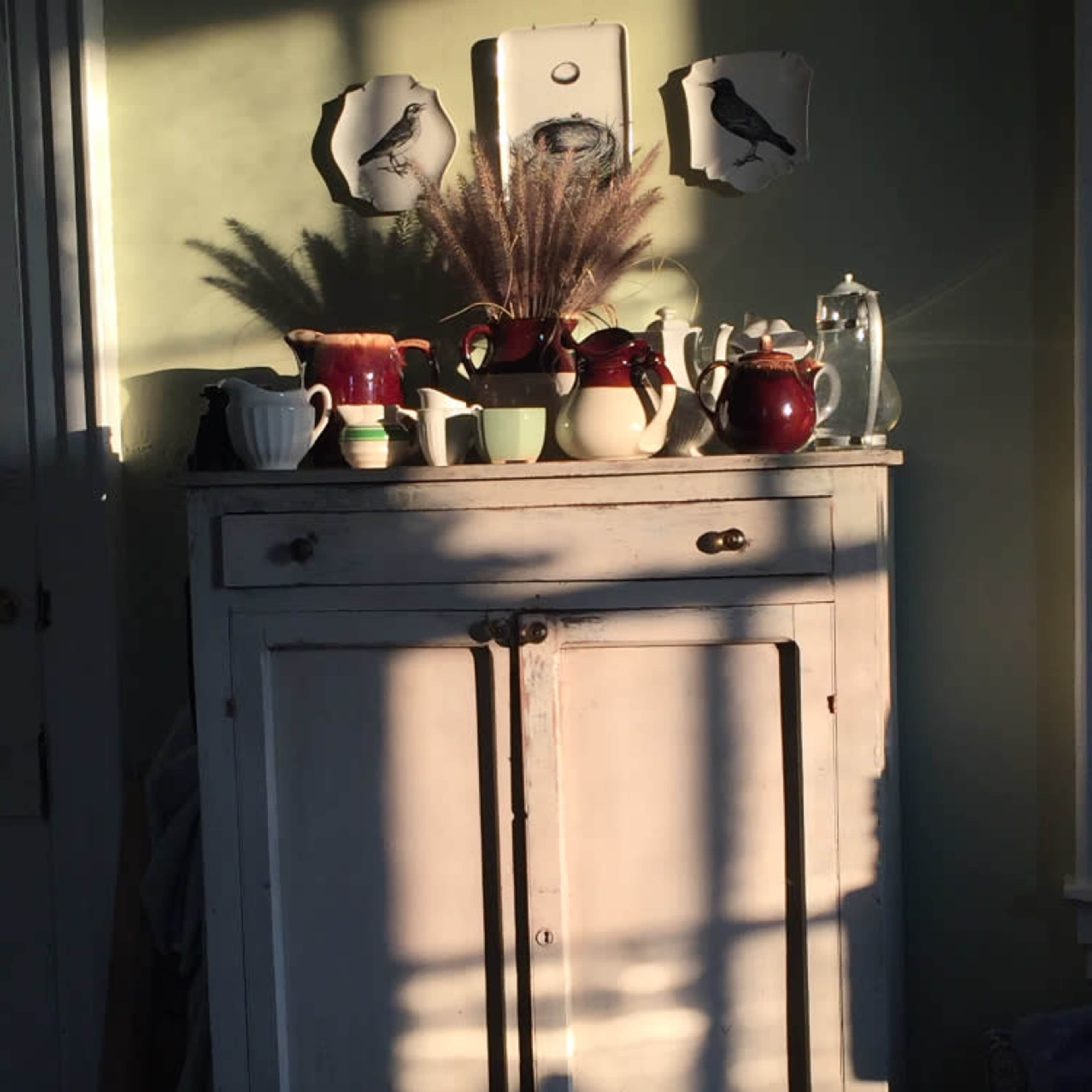 A white wooden cabinet displays various ceramic teapots and a pitcher along with decorative wall plates, illuminated by soft sunlight casting shadows across the surface.