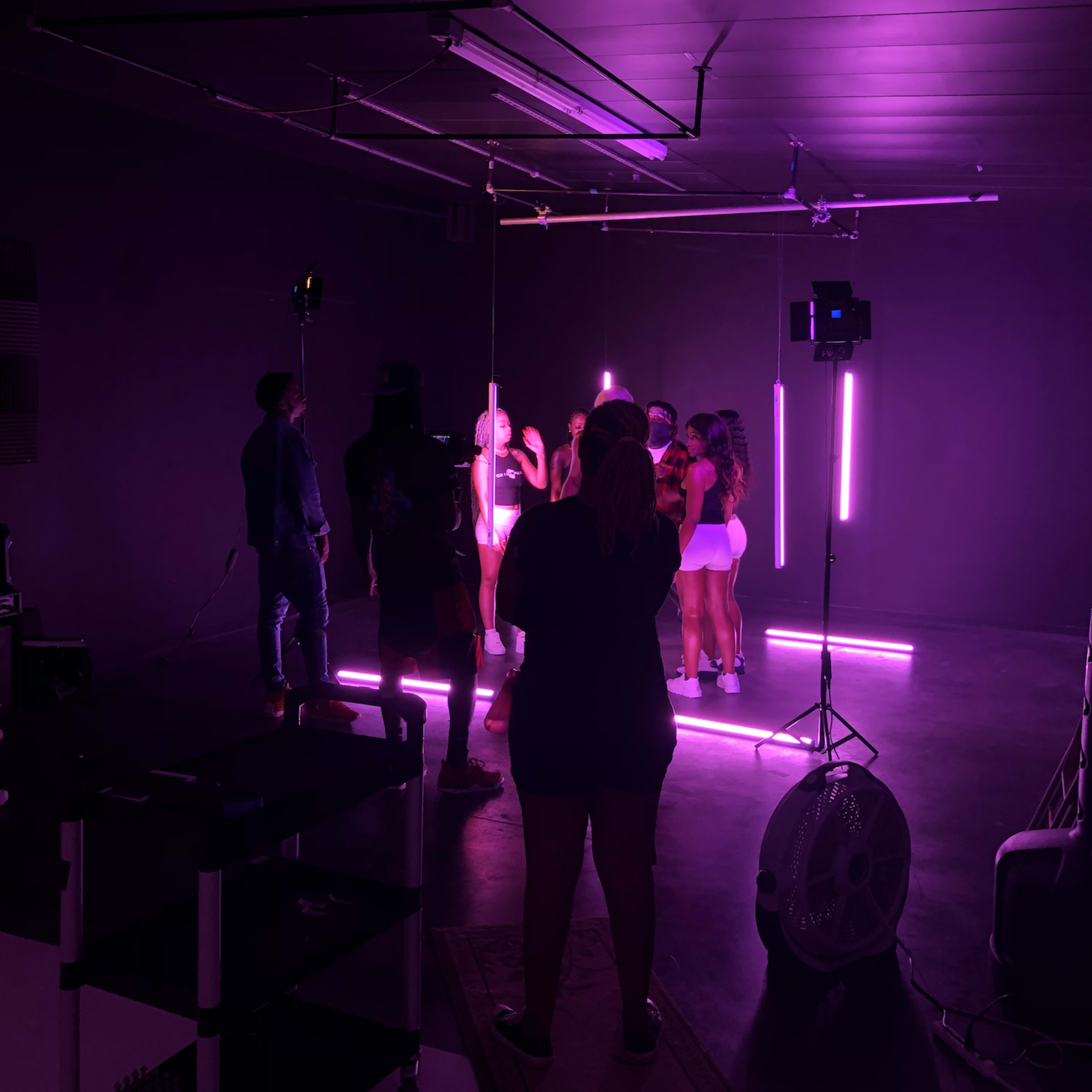 A group of people is gathered under neon lights in a studio setting, with a fan and equipment visible in the foreground.