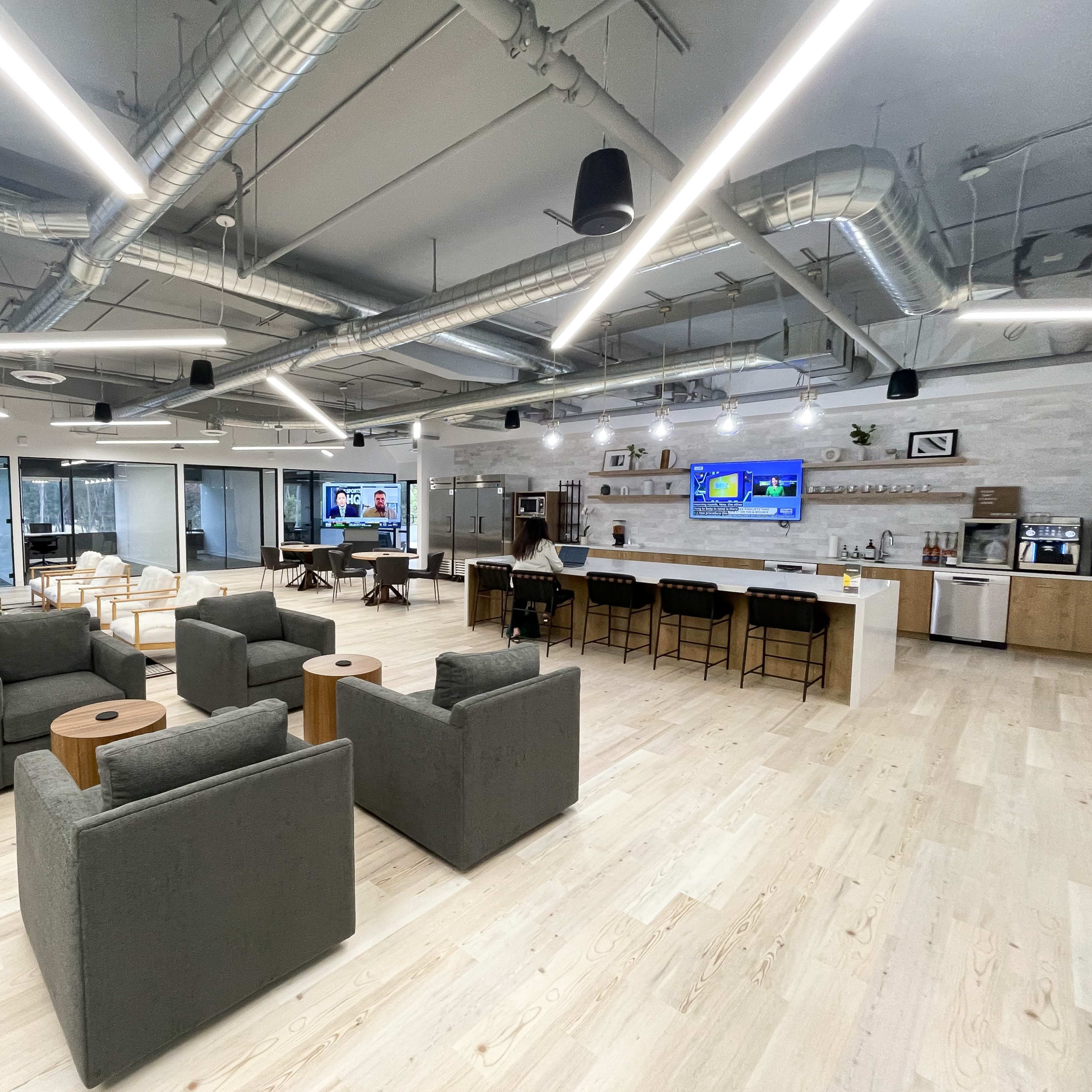 A modern office space with a lounge area featuring gray sofas and wooden tables, a kitchen area, and large windows letting in natural light.