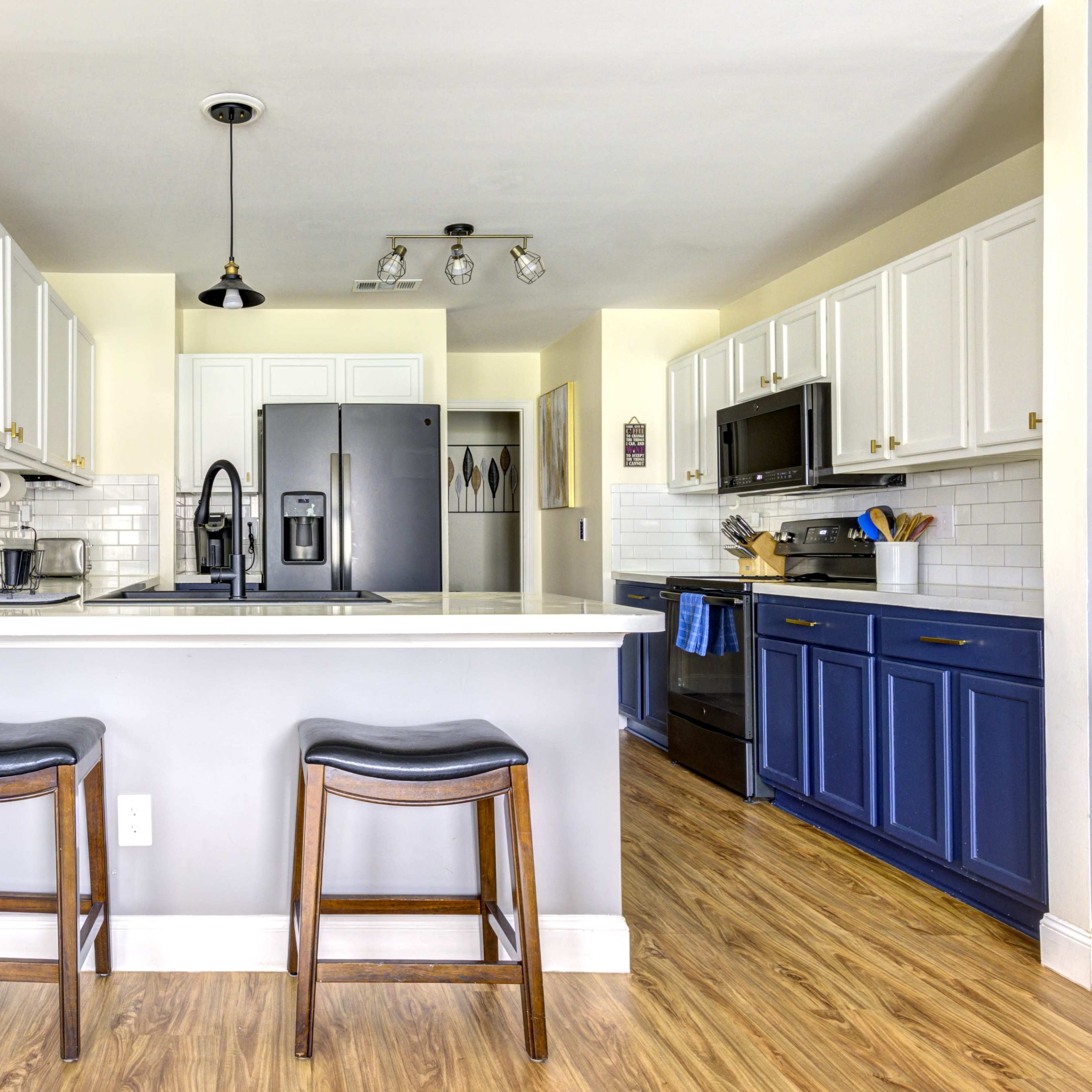 A modern kitchen features white and blue cabinetry, stainless steel appliances, and a breakfast bar with two stools.