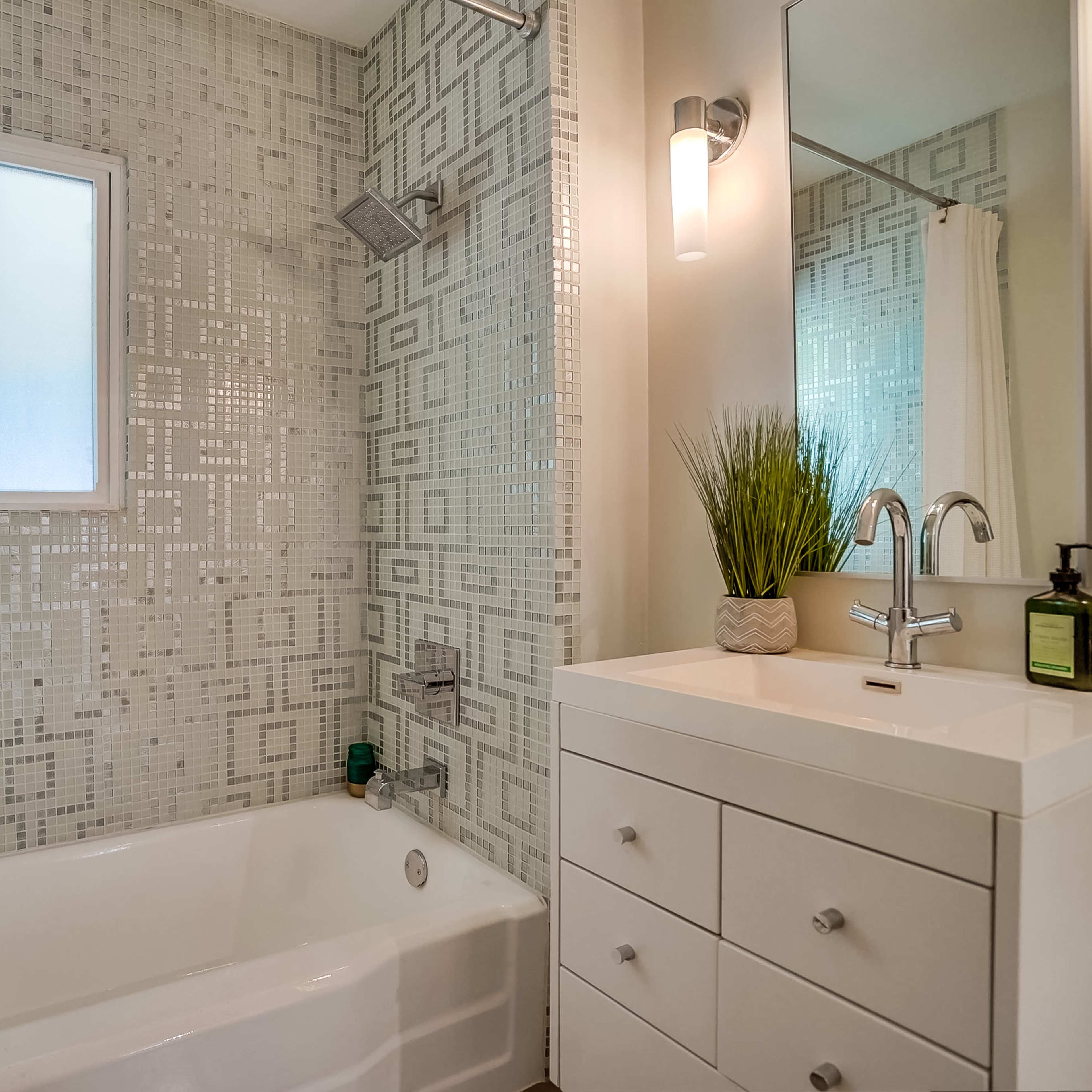 A modern bathroom with a white bathtub, a tiled wall featuring a geometric pattern, and a sink with storage drawers and a mirror.