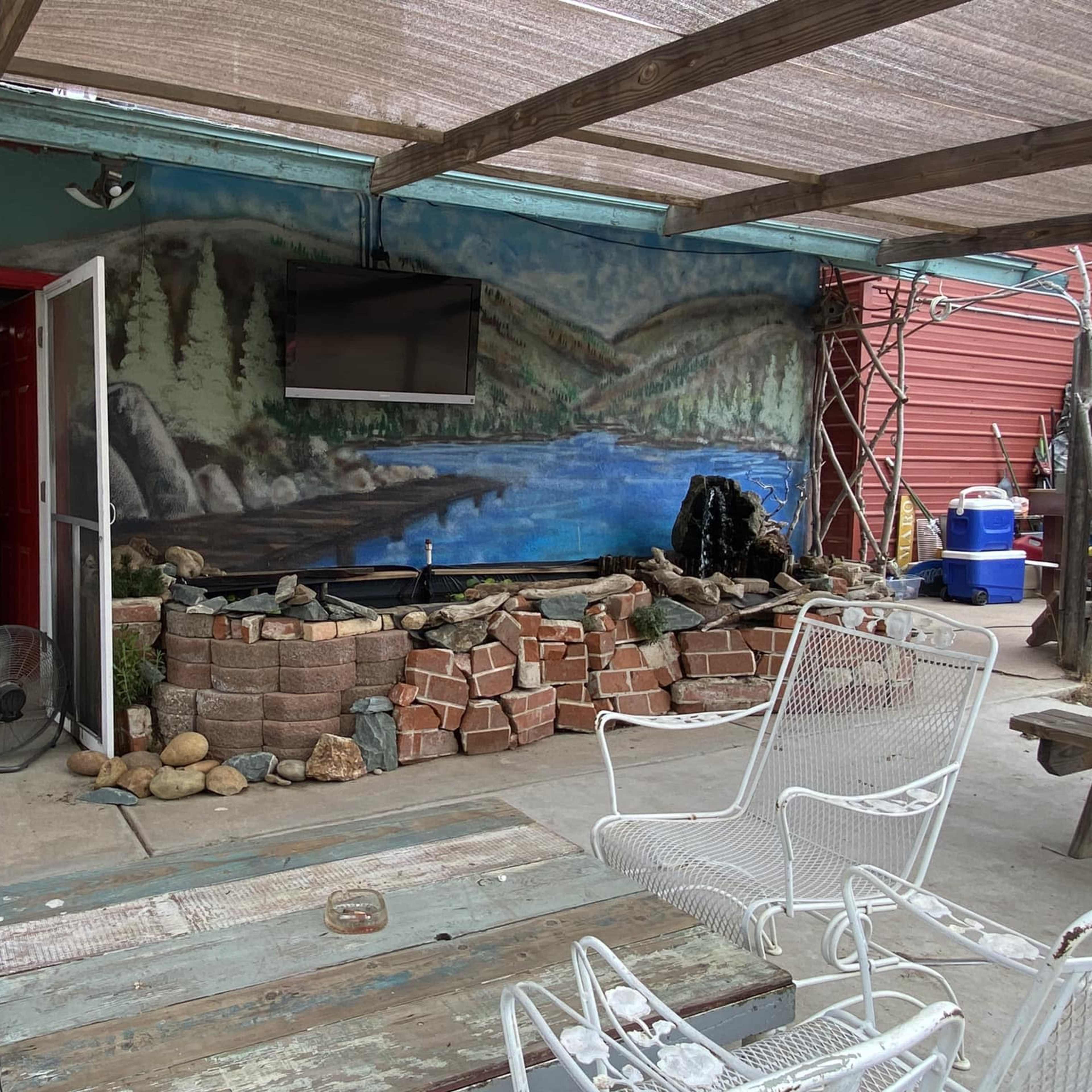 The scene shows a seating area with white chairs in front of a wall featuring a mural of a lake and mountains, alongside a decorative stone arrangement.