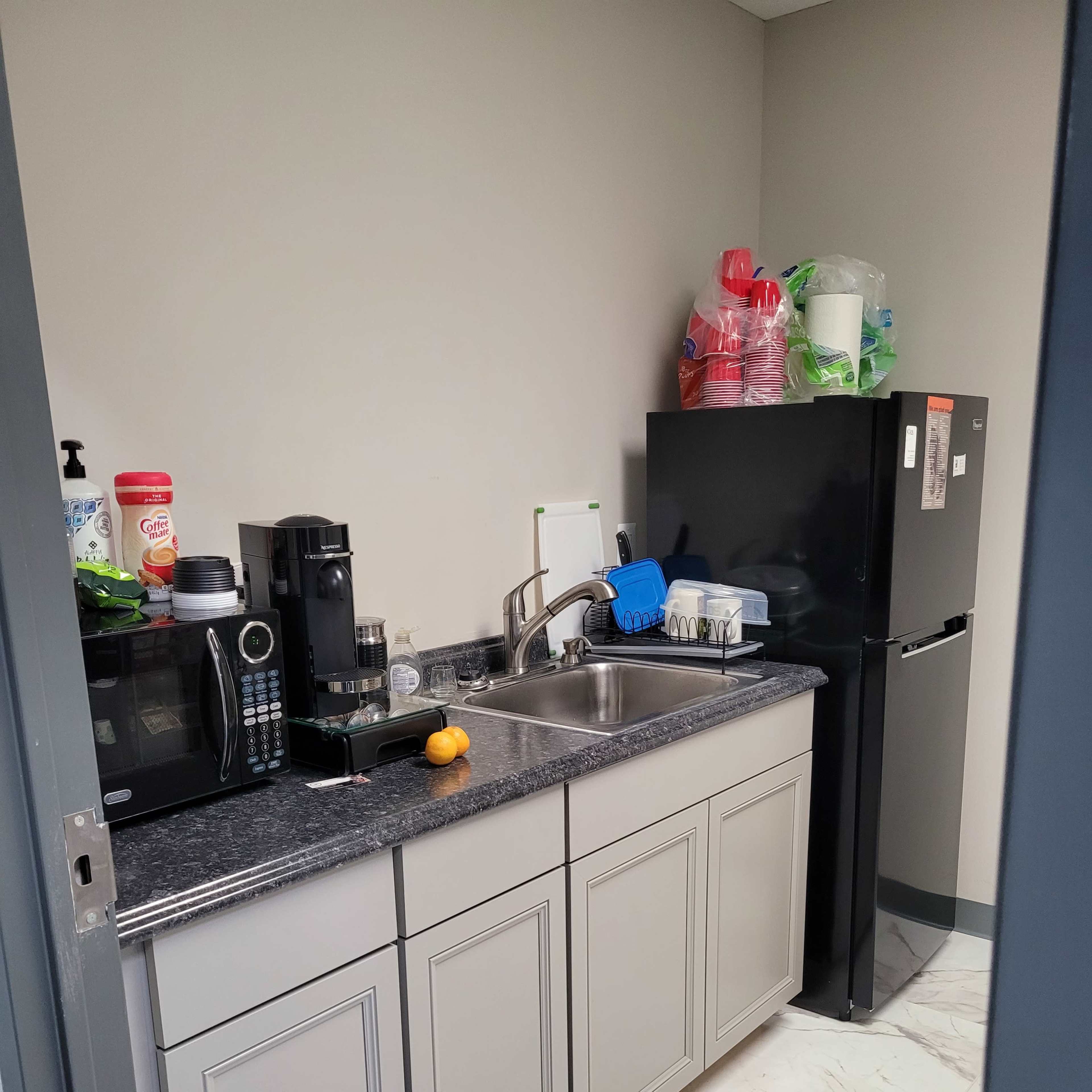 A small kitchen area with a countertop, sink, microwave, coffee maker, and a black refrigerator against a wall.