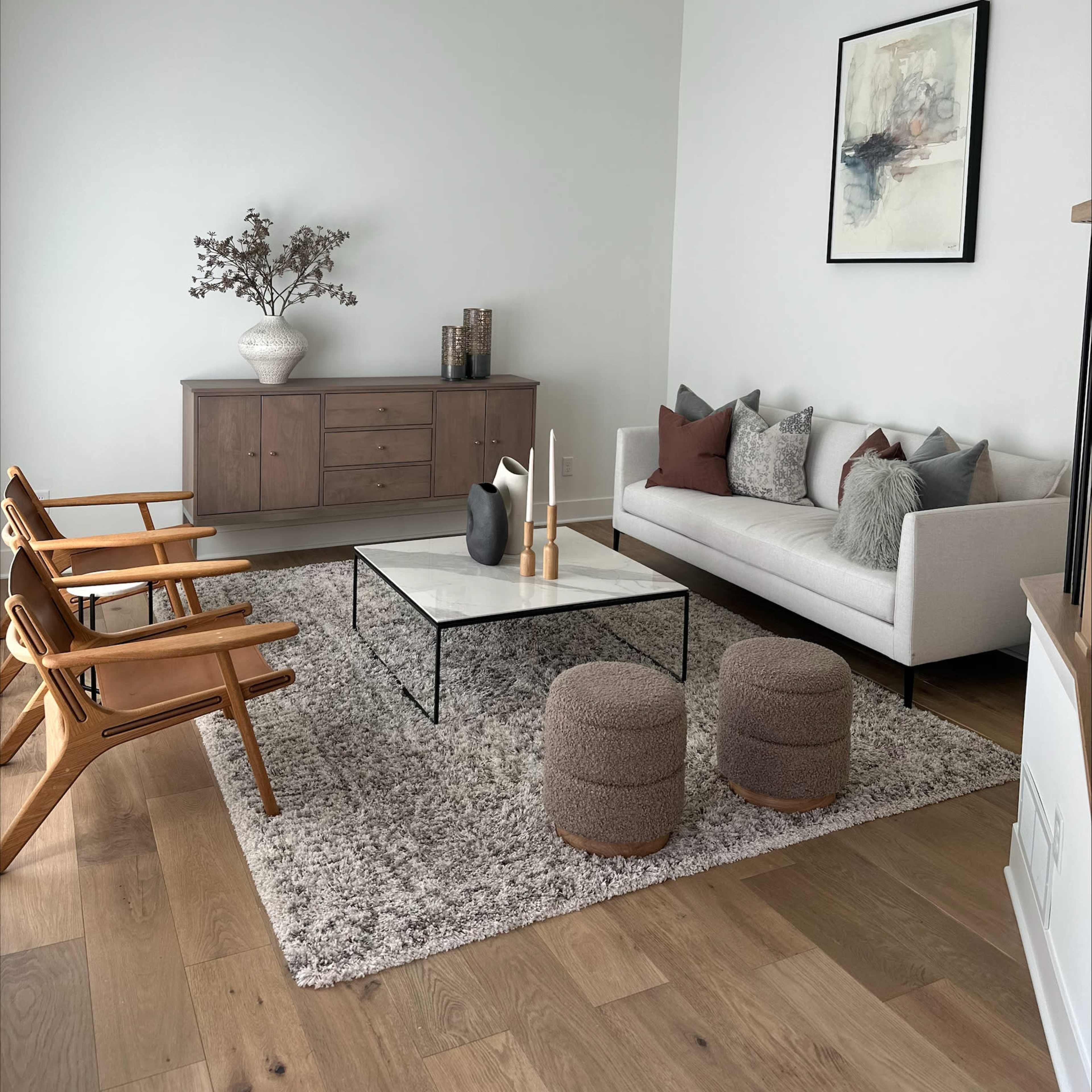A modern living room featuring a light-colored wall, a gray rug, a coffee table, a couch with decorative pillows, a wooden sideboard, and three wooden chairs.