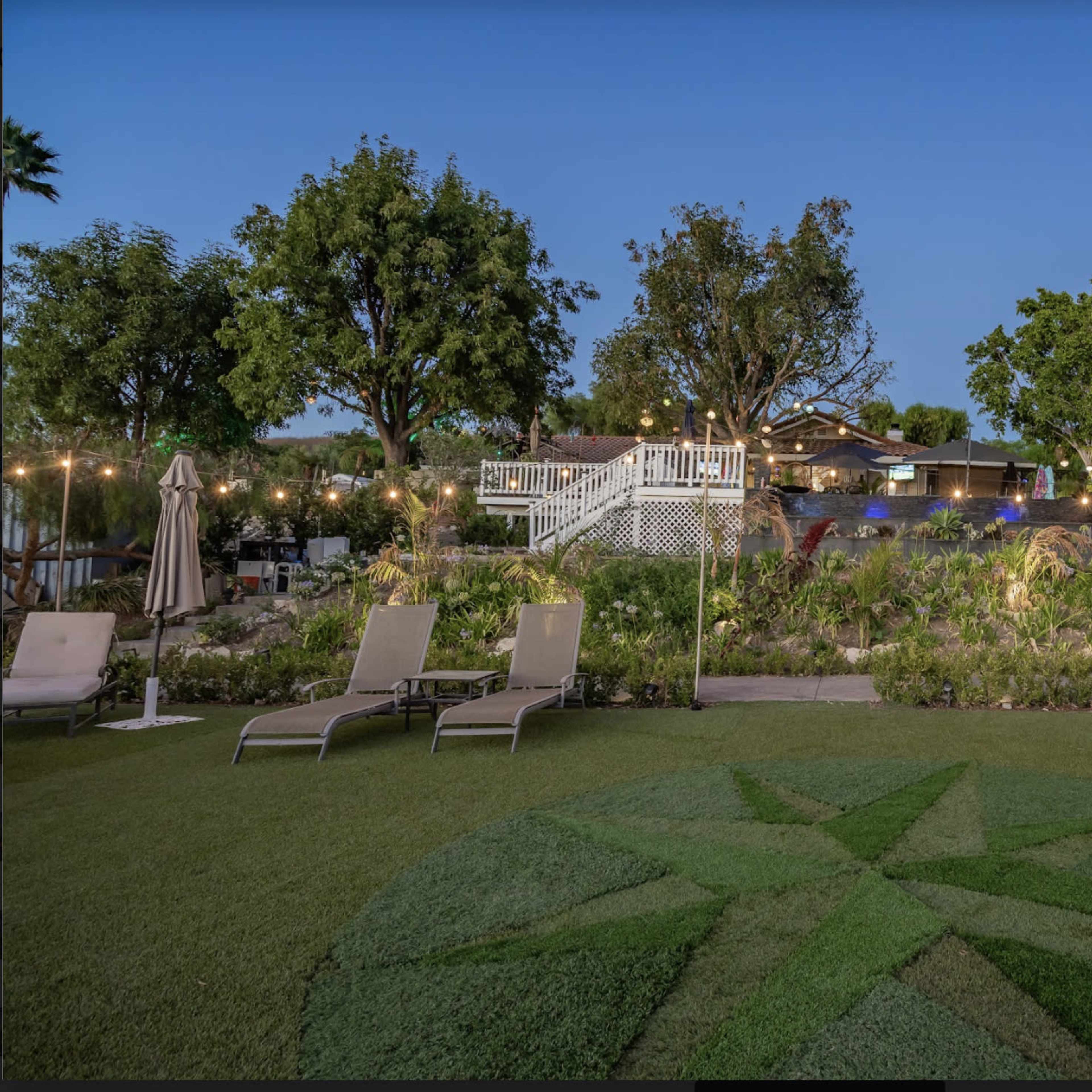 A landscaped garden area with a star-shaped design on the grass, lounge chairs, and a staircase leading to a house illuminated by evening lights.