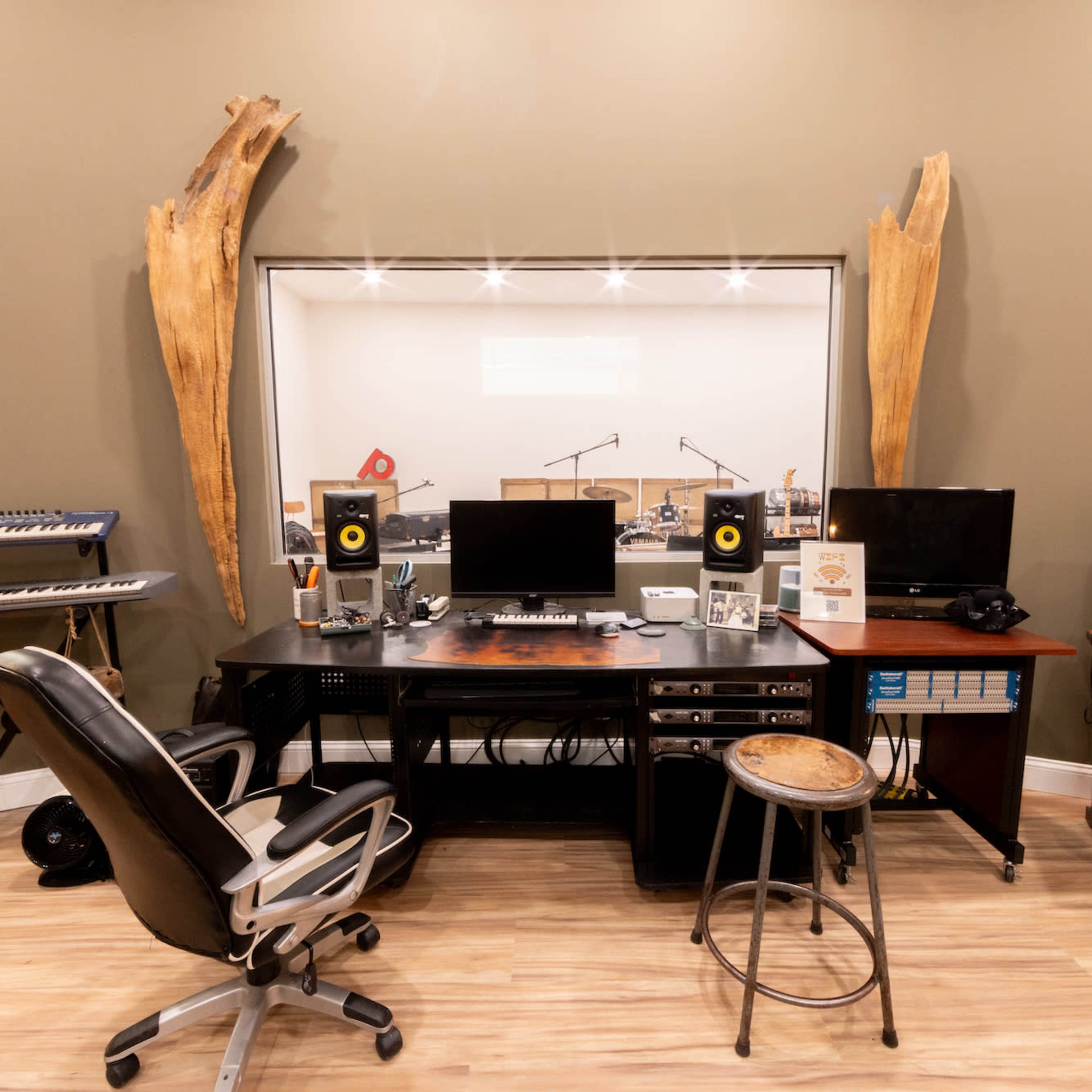 A recording studio setup features a central desk with computer monitors, audio equipment, and a chair, alongside musical instruments and a soundproof booth in the background.