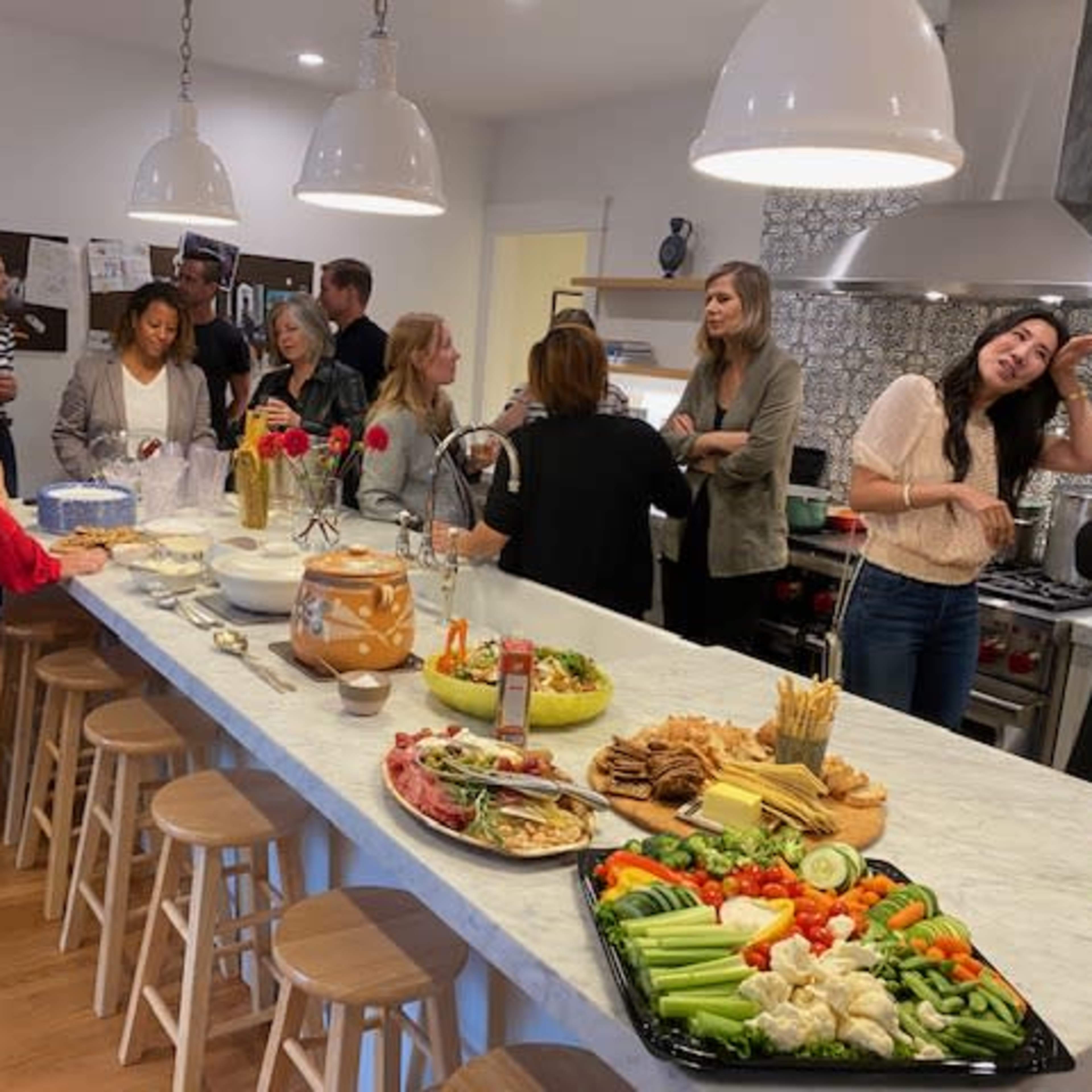 A group of people gathers in a modern kitchen, socializing around a large marble island adorned with various trays of vegetables, meats, and snacks.